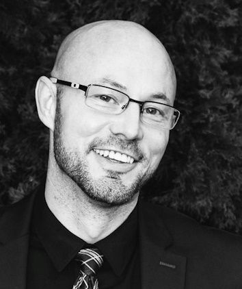 A bald man with glasses and a beard is smiling in a black and white photo.