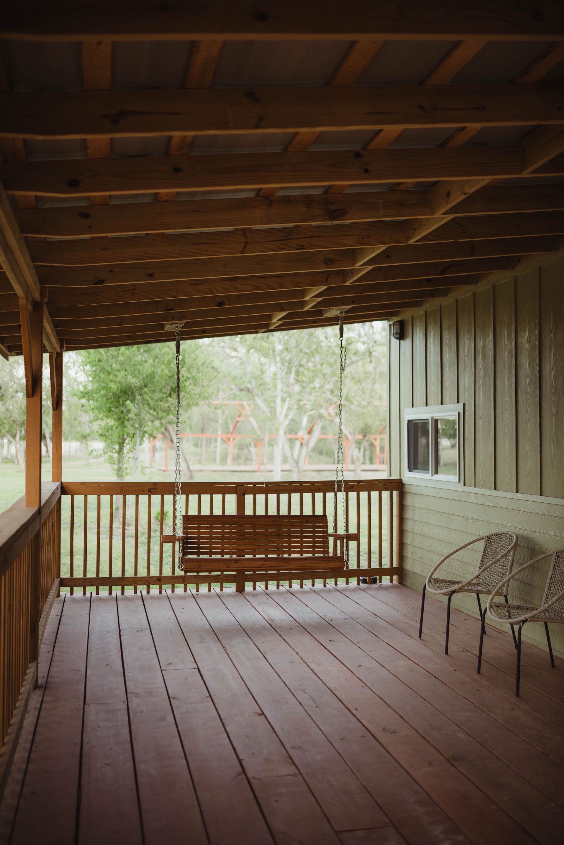 Wooden porch with a swing, railings, and chairs. Green trees are visible in the background.