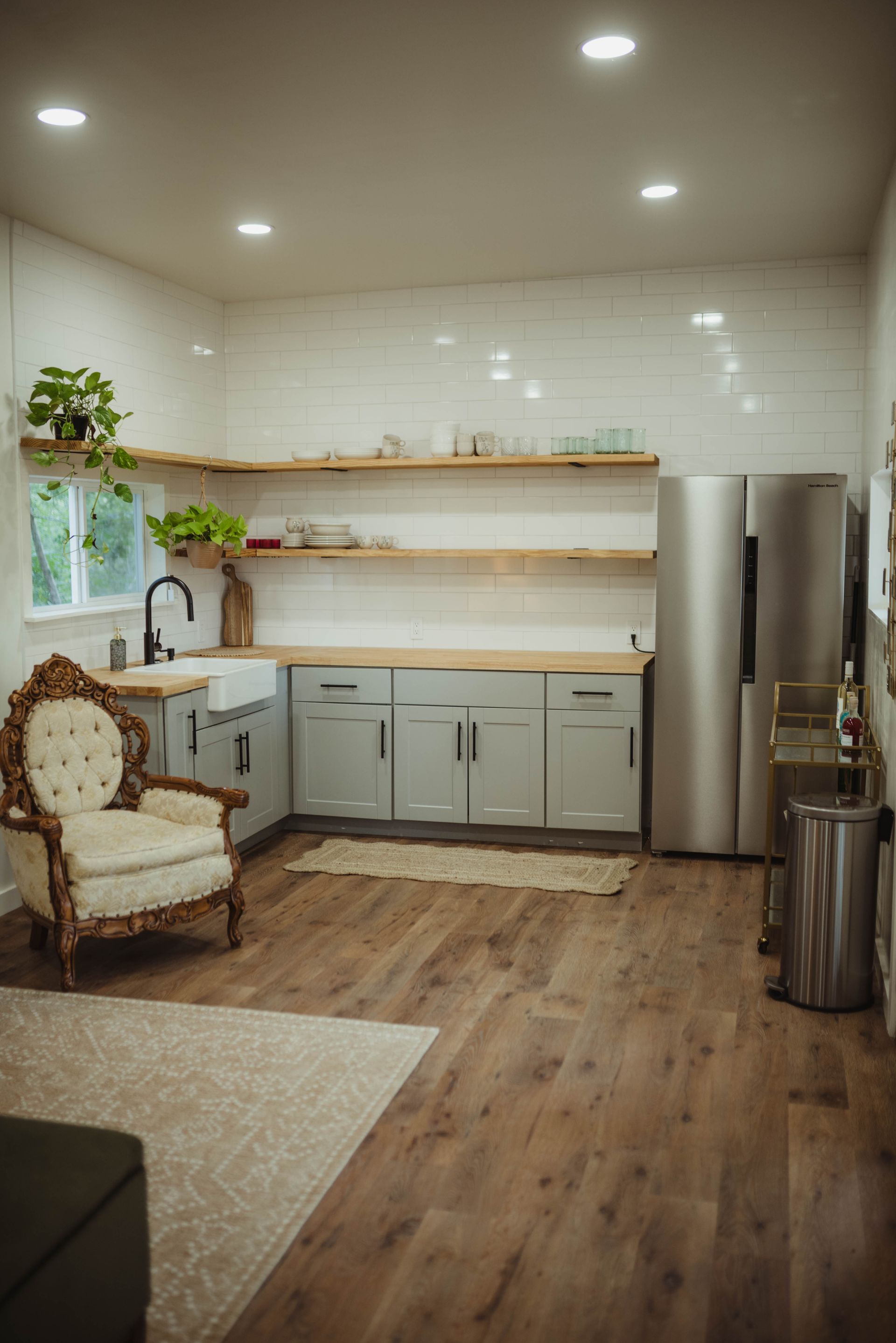 Kitchen with light wood floors, grey cabinets, open shelves, and a vintage armchair.