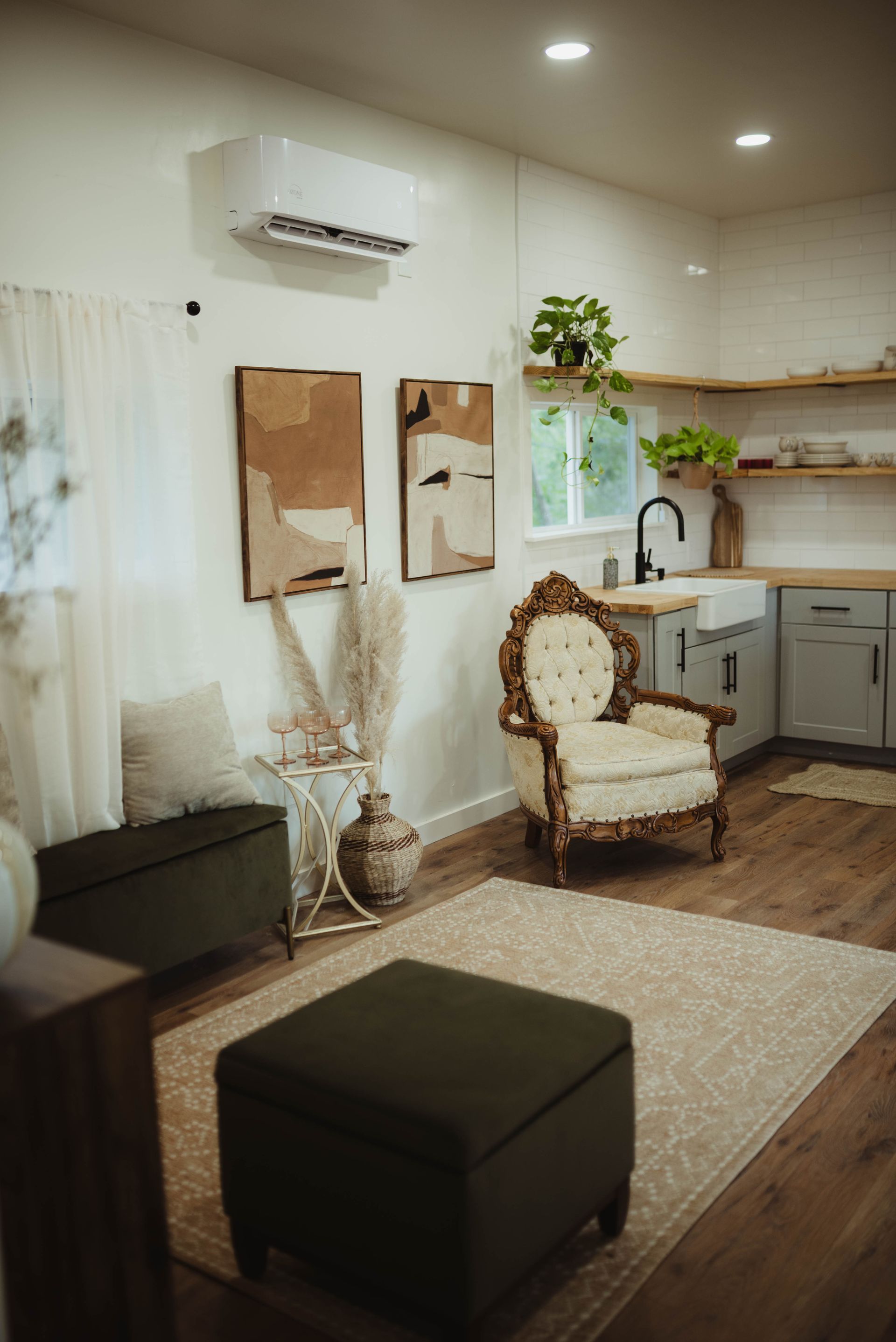 Cozy living space with armchair, ottoman, artwork, and kitchen in background; neutral tones.