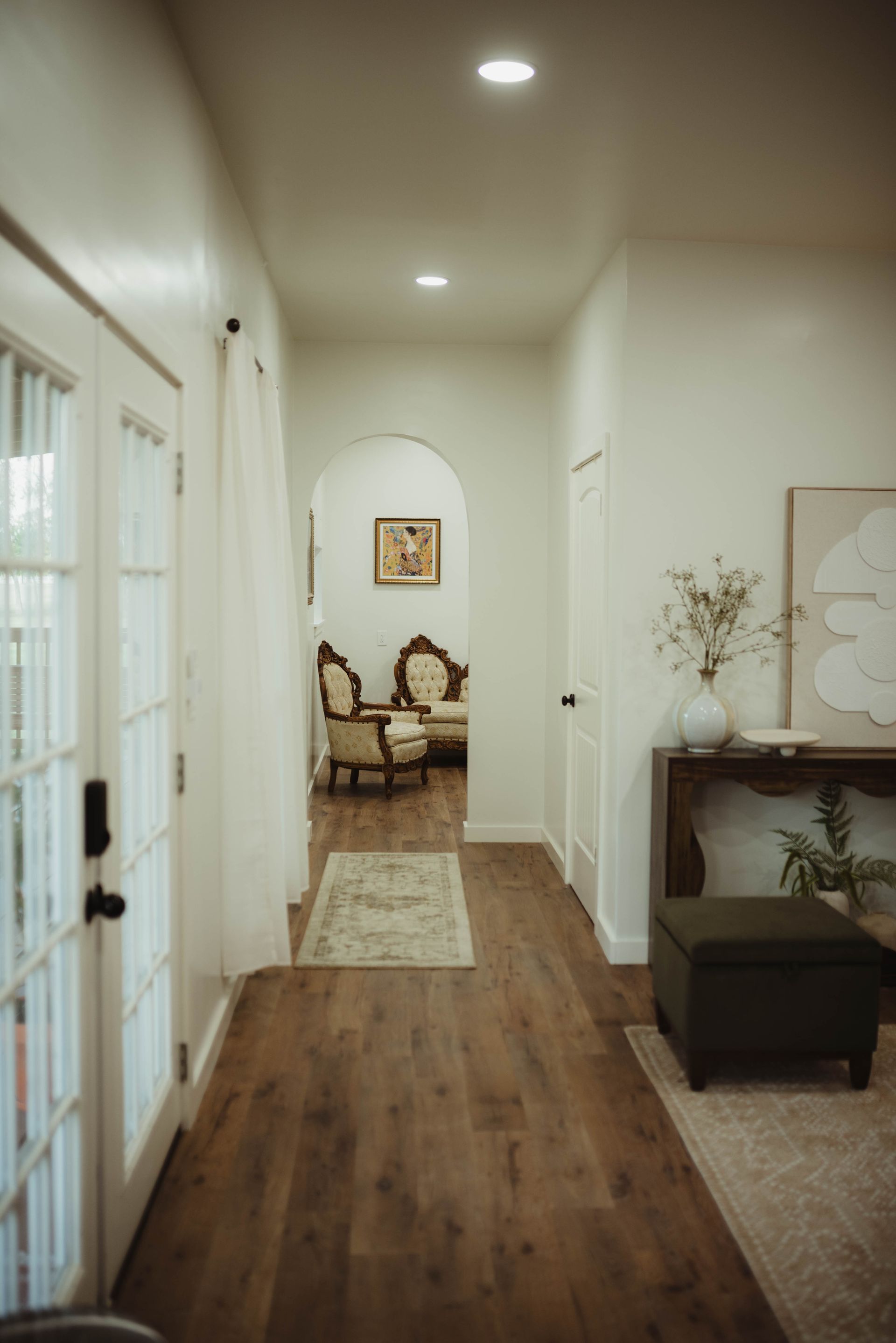 Hallway with wooden floors, white walls, and furniture in the background.