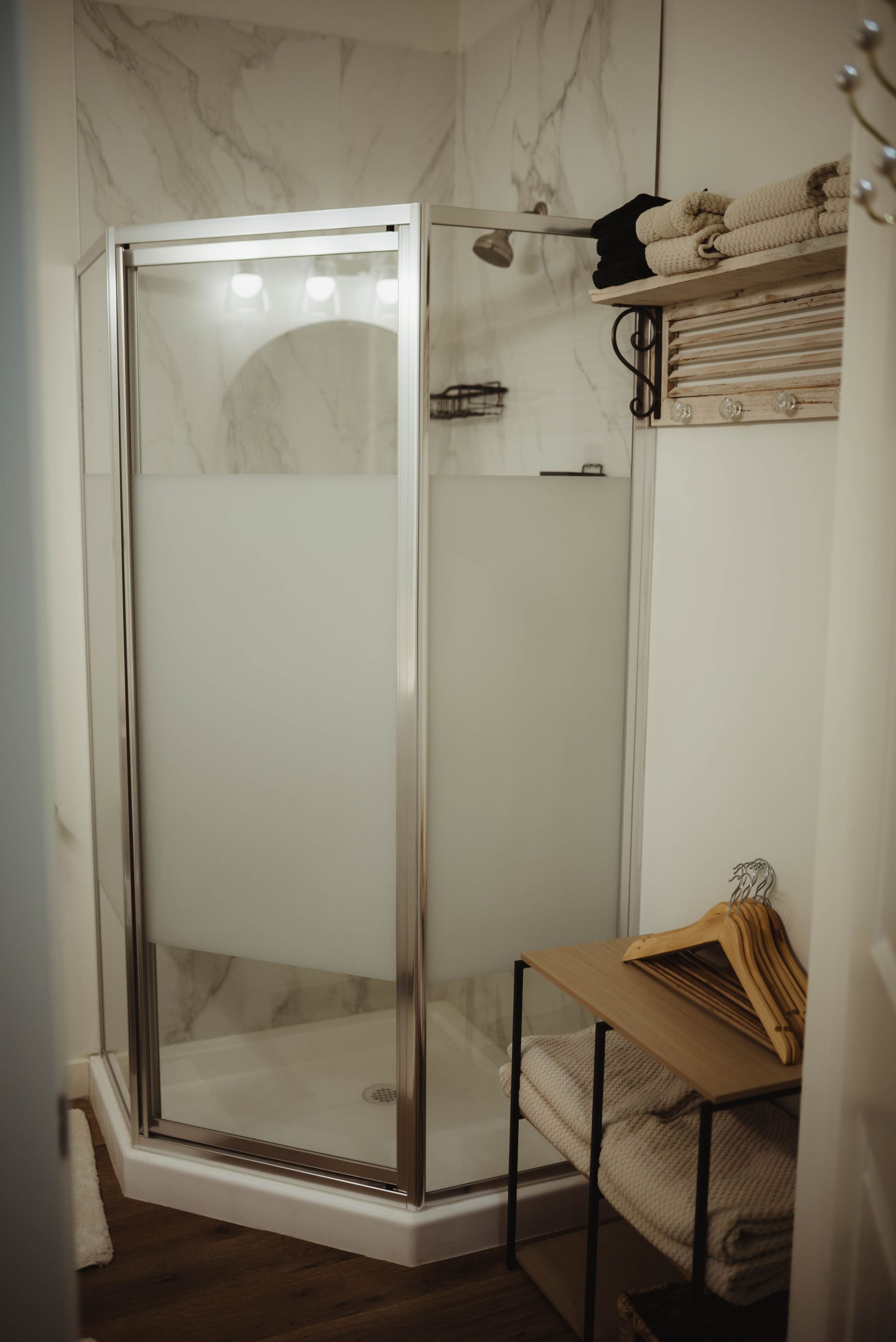 Small shower with frosted glass door, wood shelf with towels, and a small shelf with hangers.