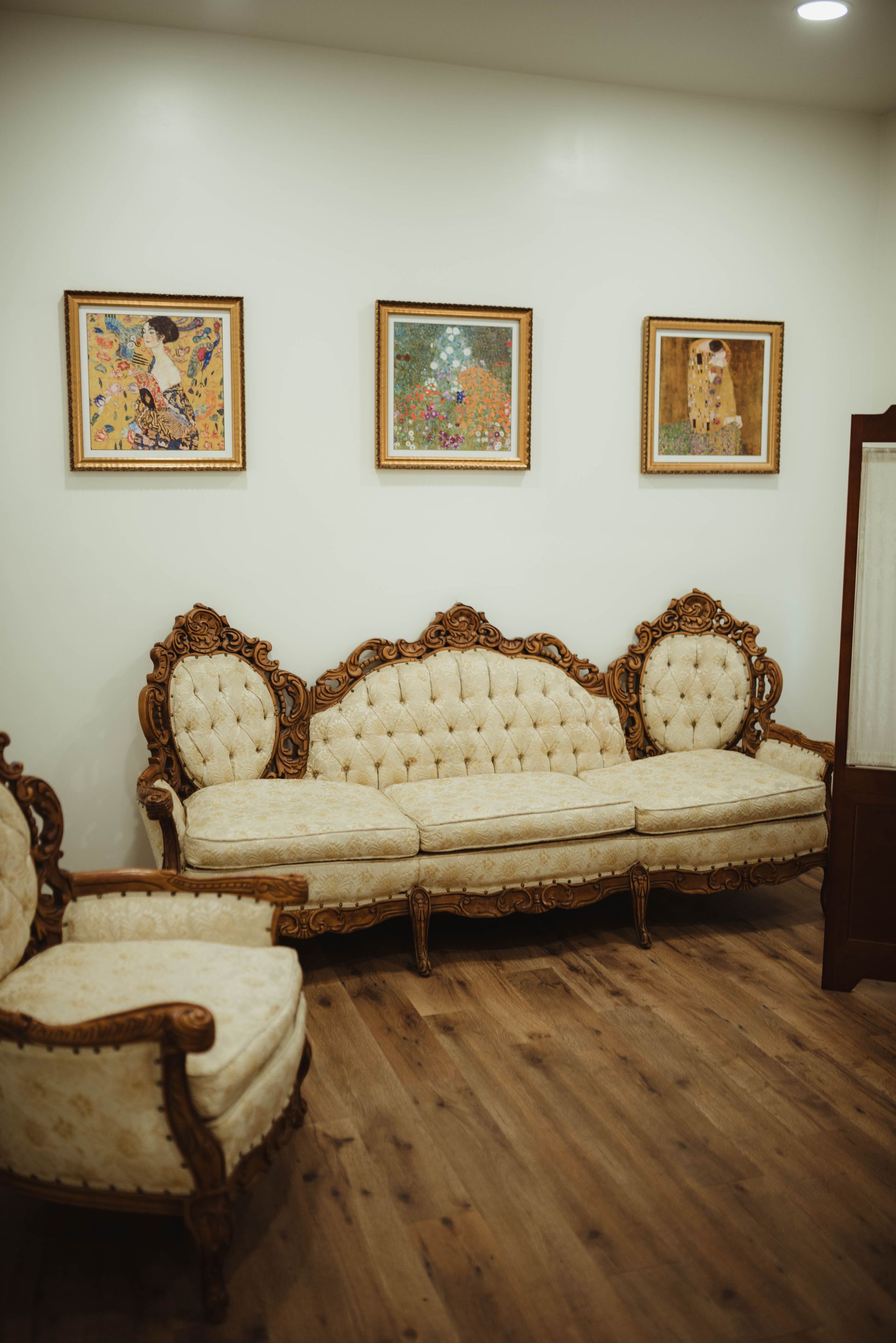 Ornate sofa and chair in a room with three framed paintings on the wall and hardwood floors.
