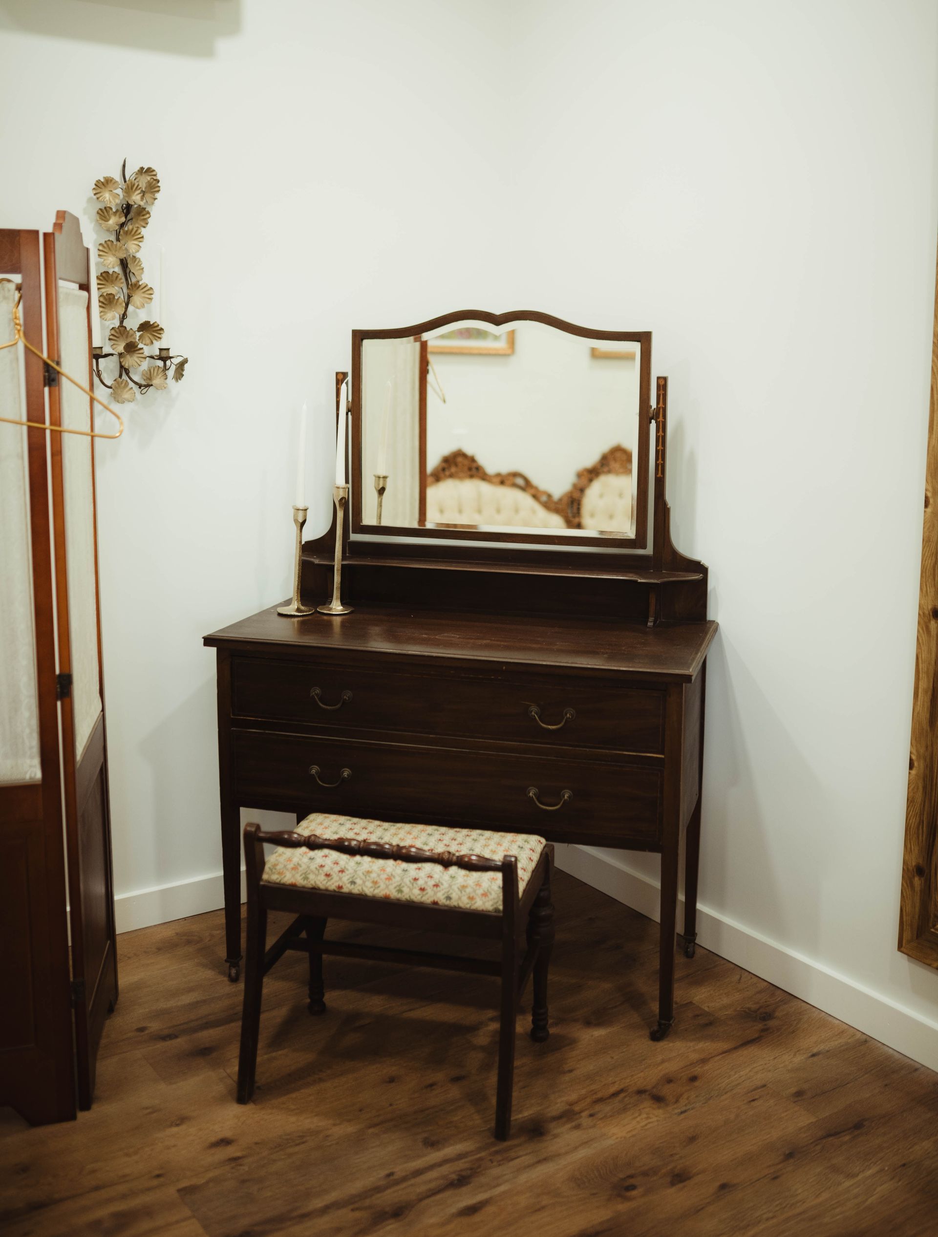 Dark wood vanity with a trifold mirror, stool, and a candle, set in a corner.
