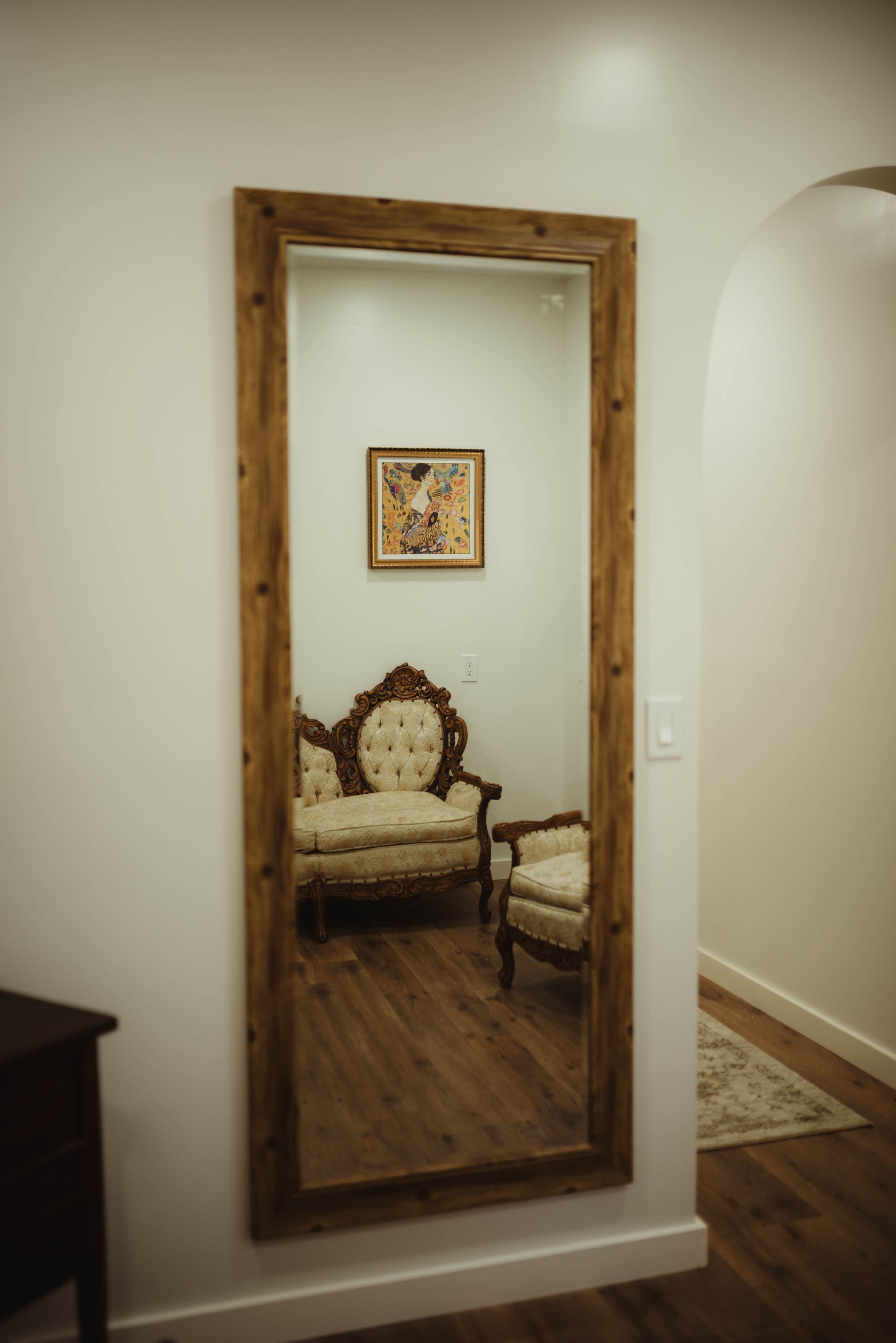 Full-length mirror on a white wall reflecting a sofa, chair, and artwork in a room with wood floors.