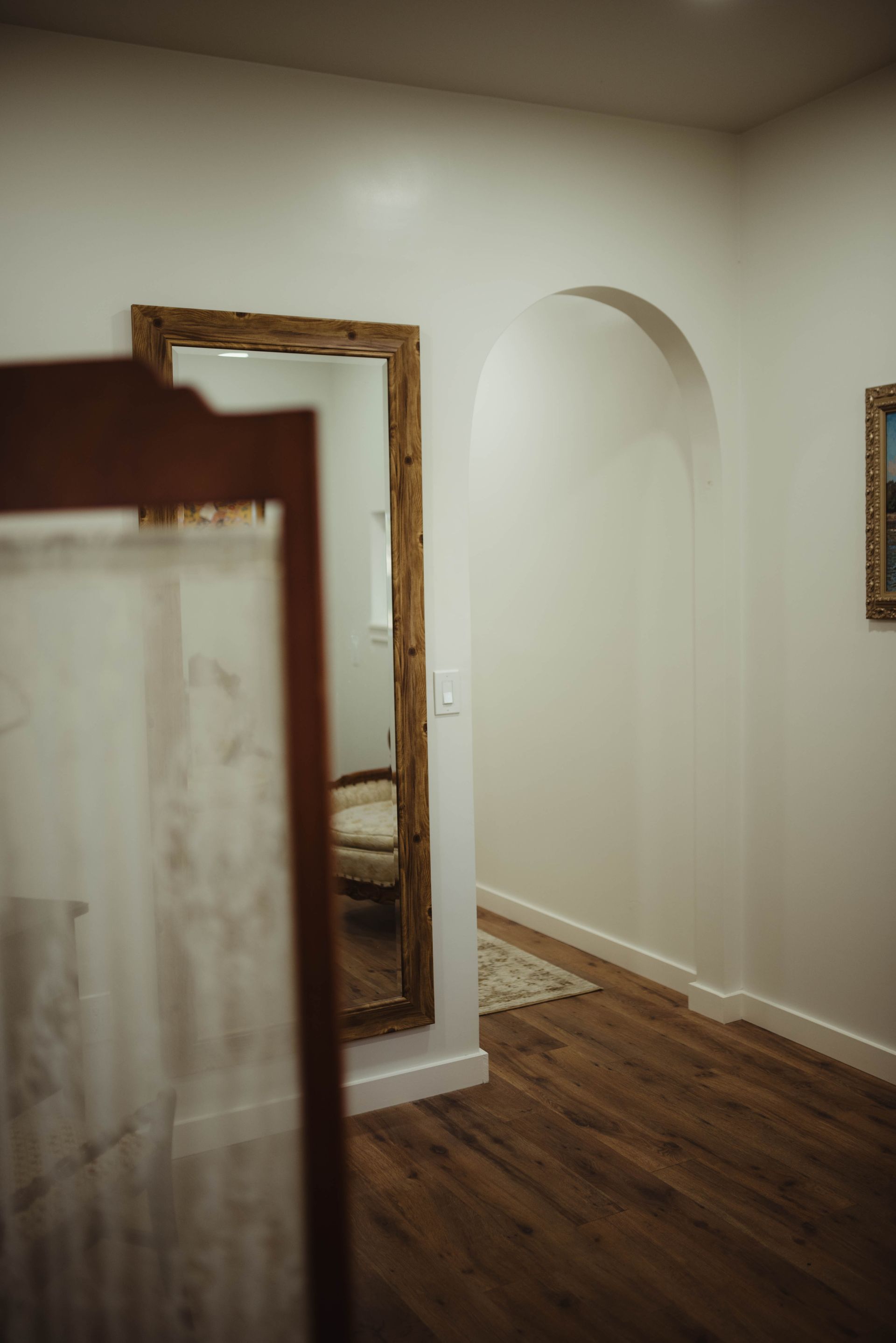 A full-length rustic mirror reflects a room with arched doorway, wood floor, and white walls.