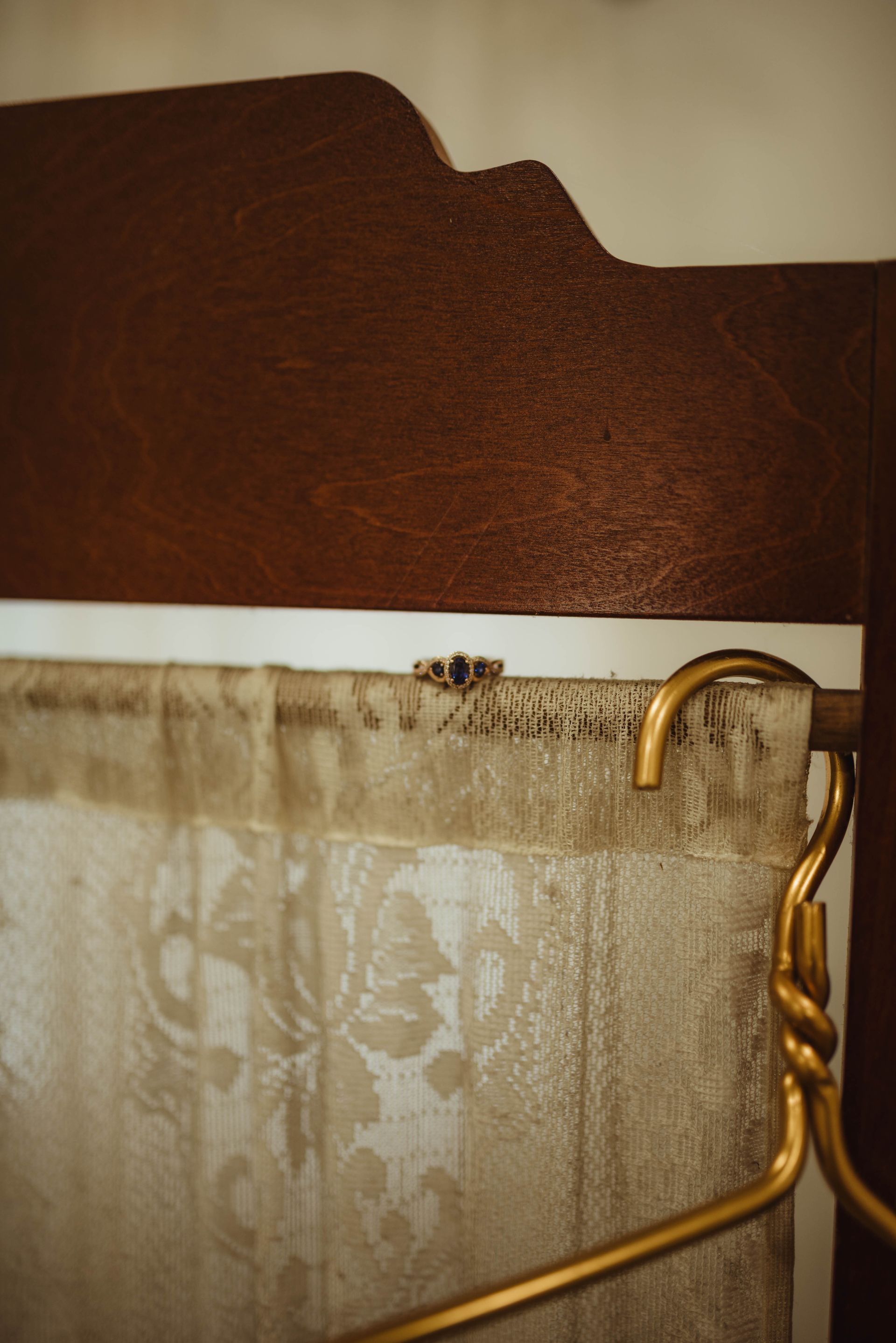 Wedding rings on a lace curtain rod with a gold hanger, set against a wooden headboard.