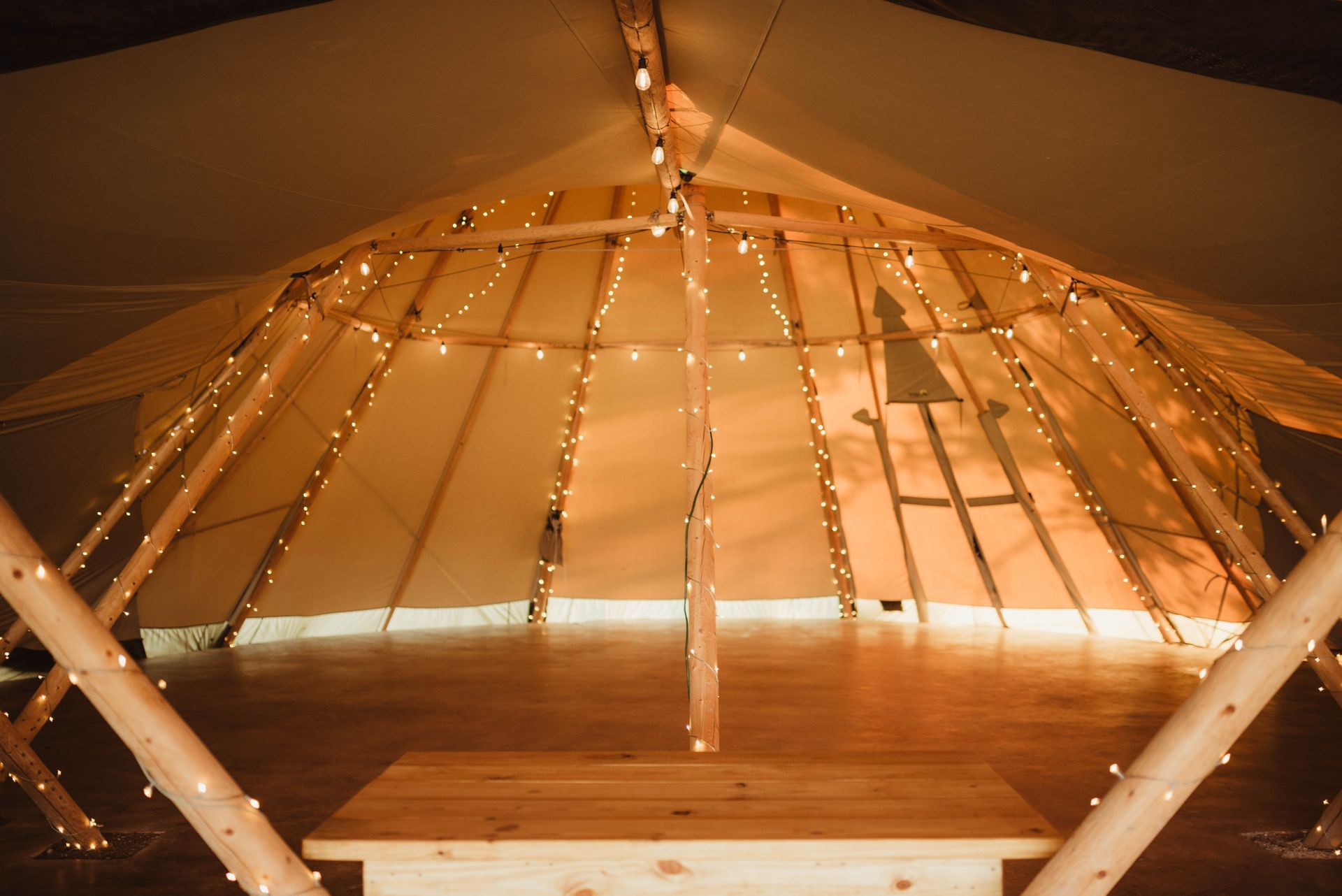 Inside a warm lit teepee, a wooden table sits on a wood floor, fairy lights draped.