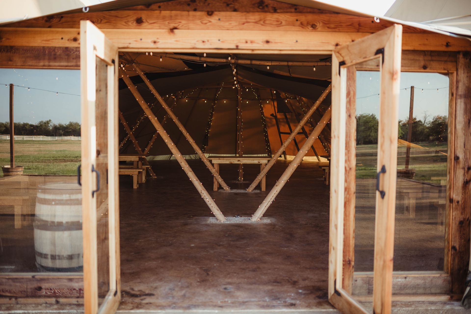 Wooden doors opening to a teepee-style tent; a barrel sits beside the entrance, open field.