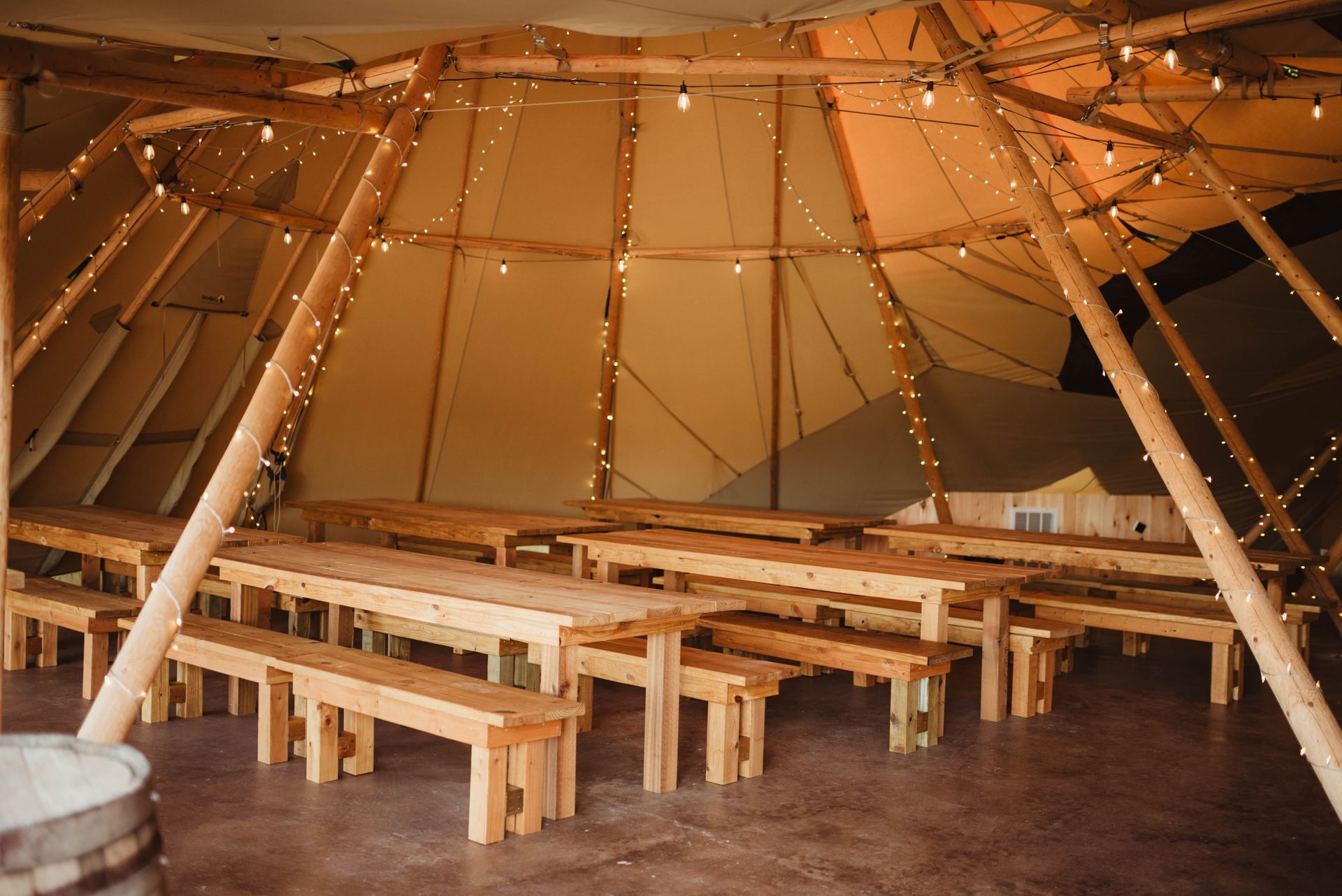Inside a large tent, wooden tables and benches are set up. String lights hang from the wooden structure.