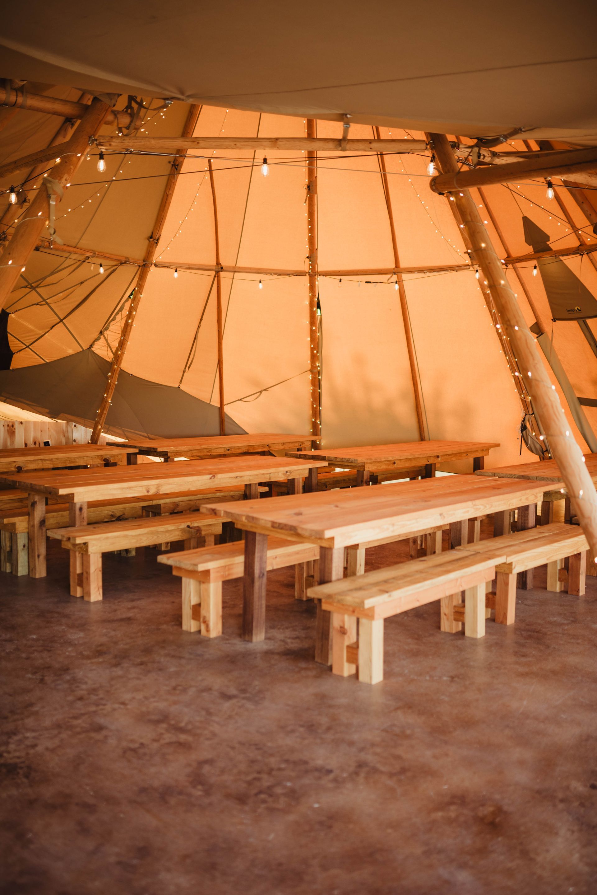 Wooden benches and tables set up inside a large teepee-style tent, lit by string lights.