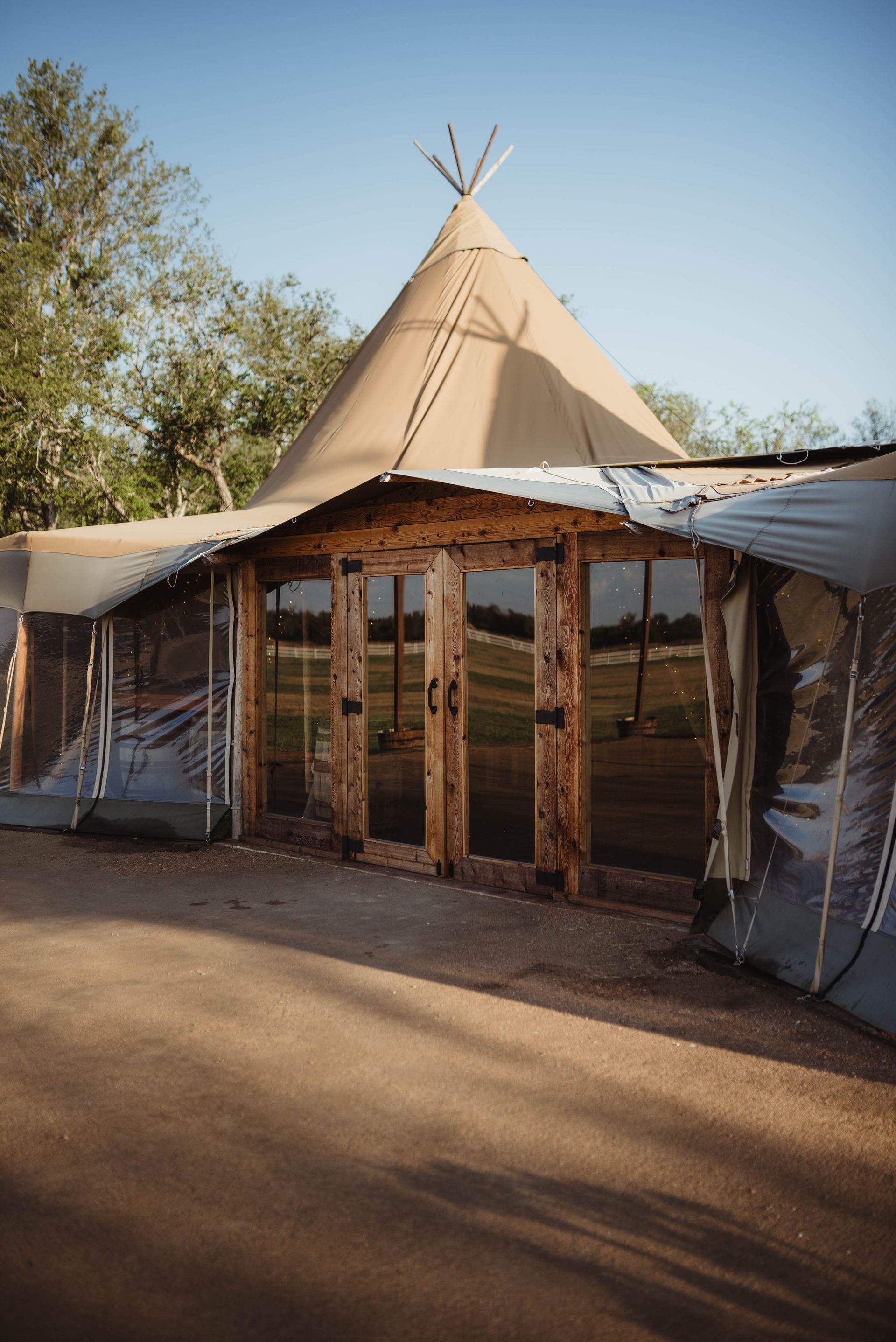 A teepee-style tent with wooden doors and clear side panels, set on a paved surface in a sunny outdoor location.