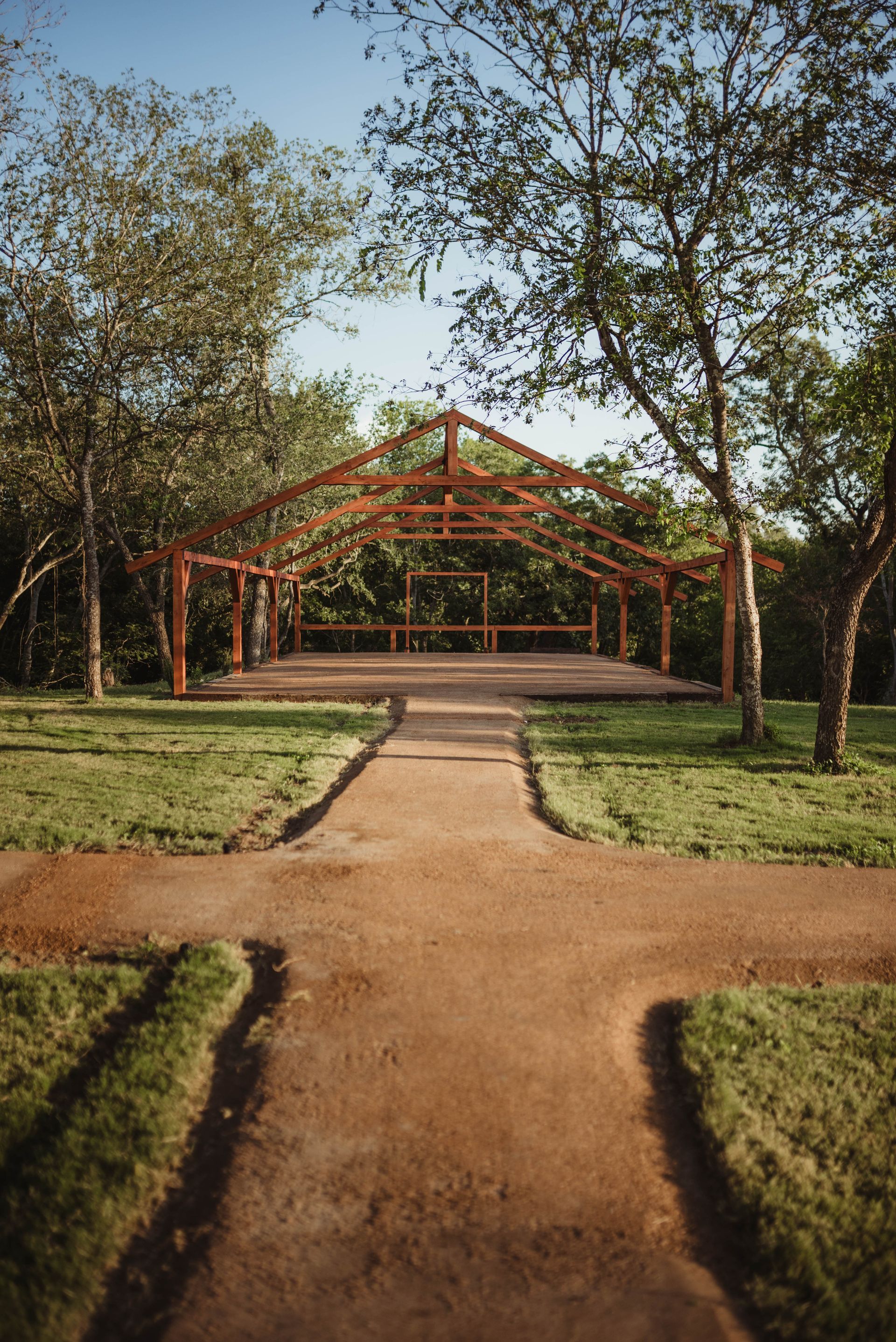 Brown dirt path leads to a wooden pavilion in a green, sunny park.