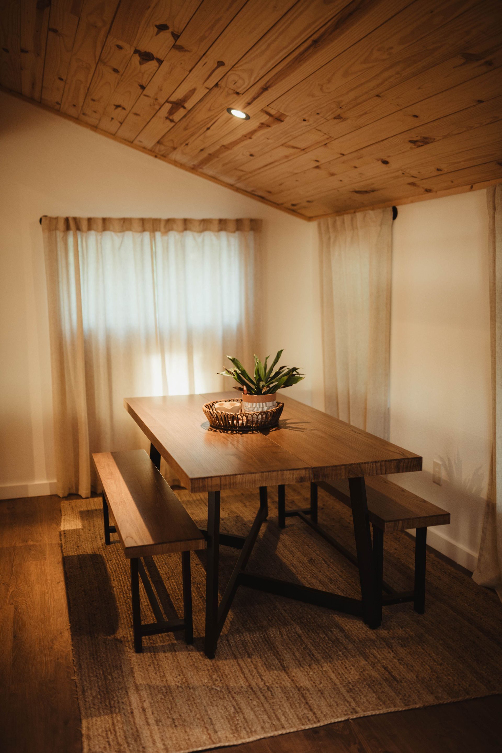 Wooden dining table and benches in a light-filled room with a wood ceiling, and a window with sheer curtains.