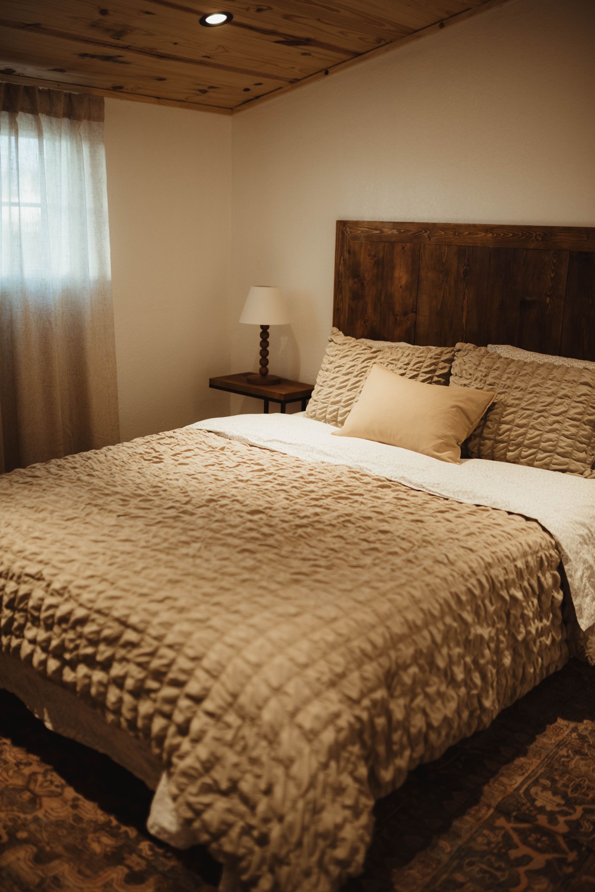 Cozy bedroom with a bed covered in tan quilted bedding, wooden headboard, and a small bedside table with lamp.