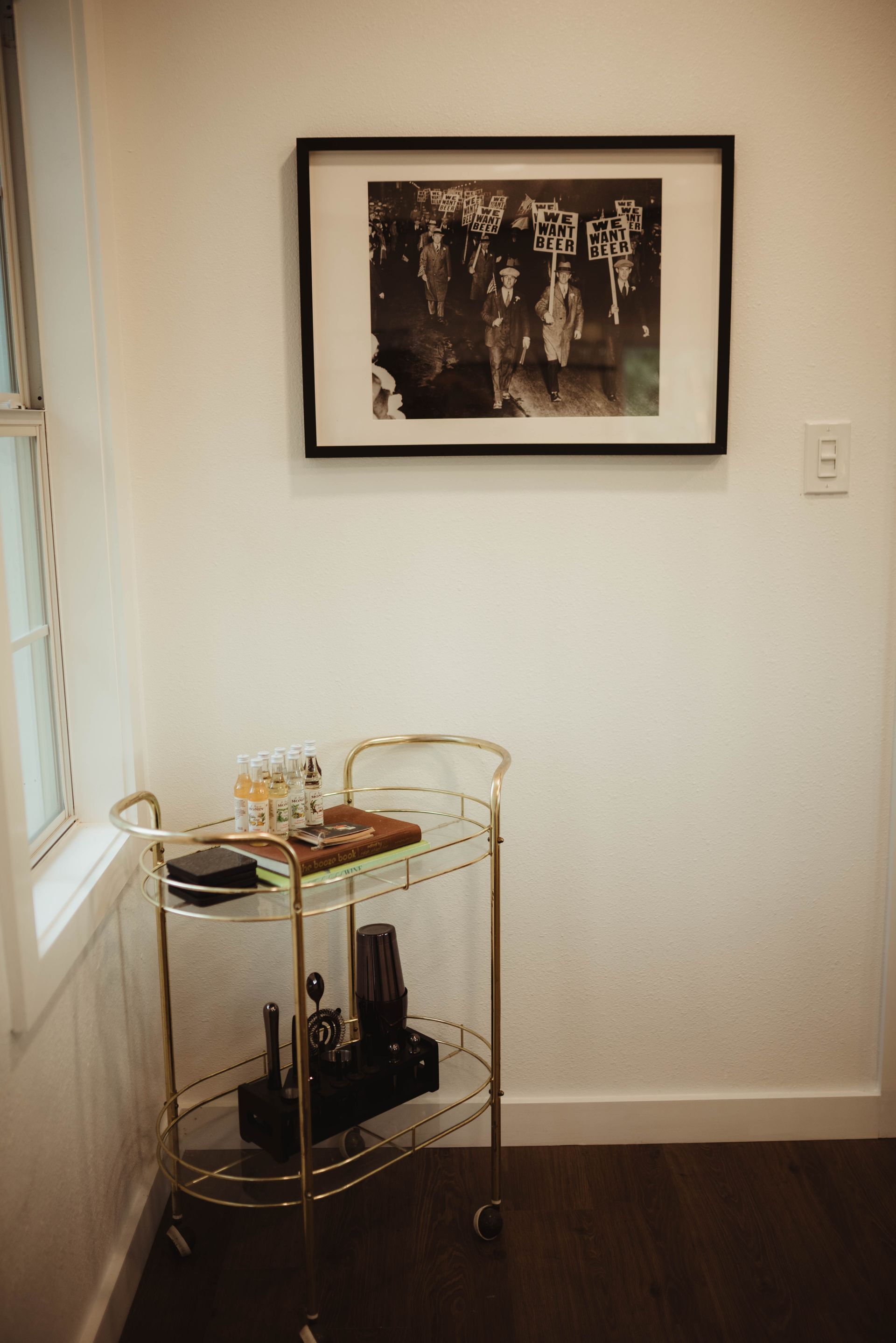 A gold bar cart with drinks sits under a framed black and white photo in a white-walled room.
