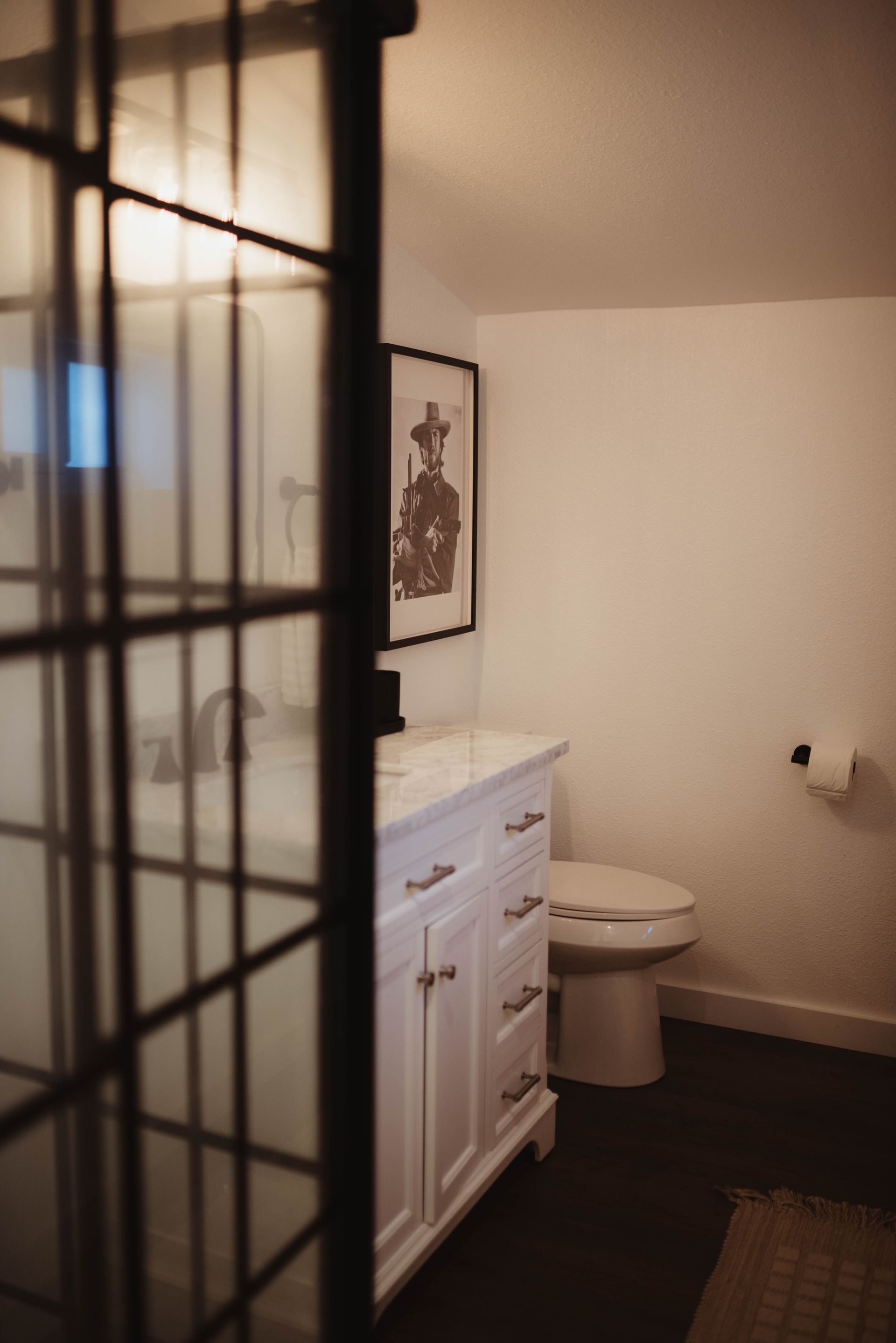 Bathroom with white vanity, toilet, and framed artwork, viewed through a glass paneled door.