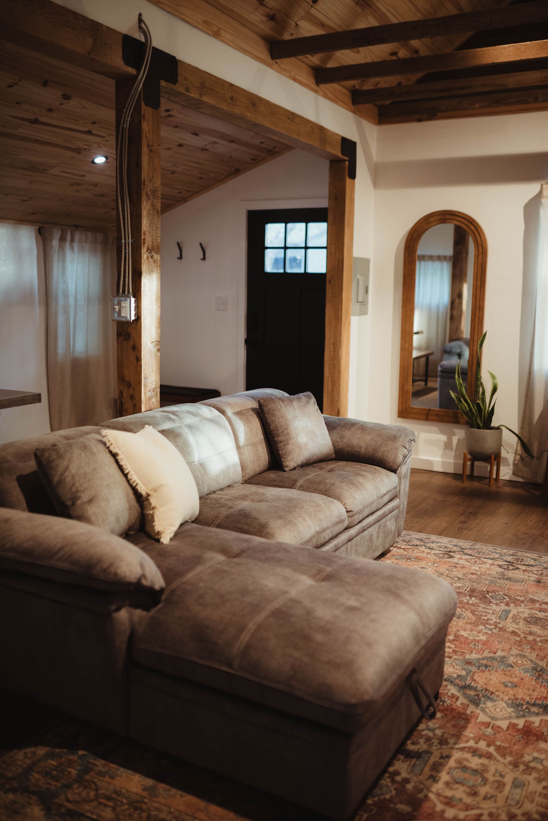 Cozy living room with a brown sectional sofa, arched mirror, and patterned rug. Wooden beams and dark door in the background.