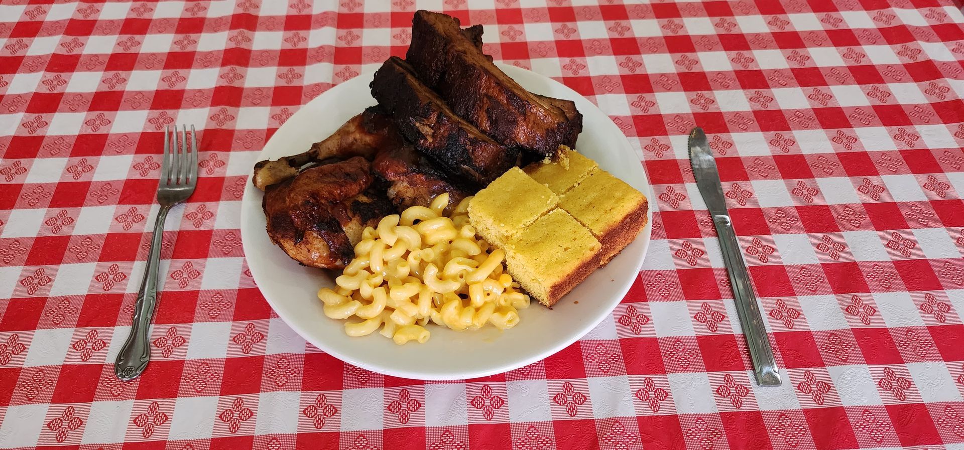 A plate of food with ribs macaroni and cornbread on a checkered table cloth.