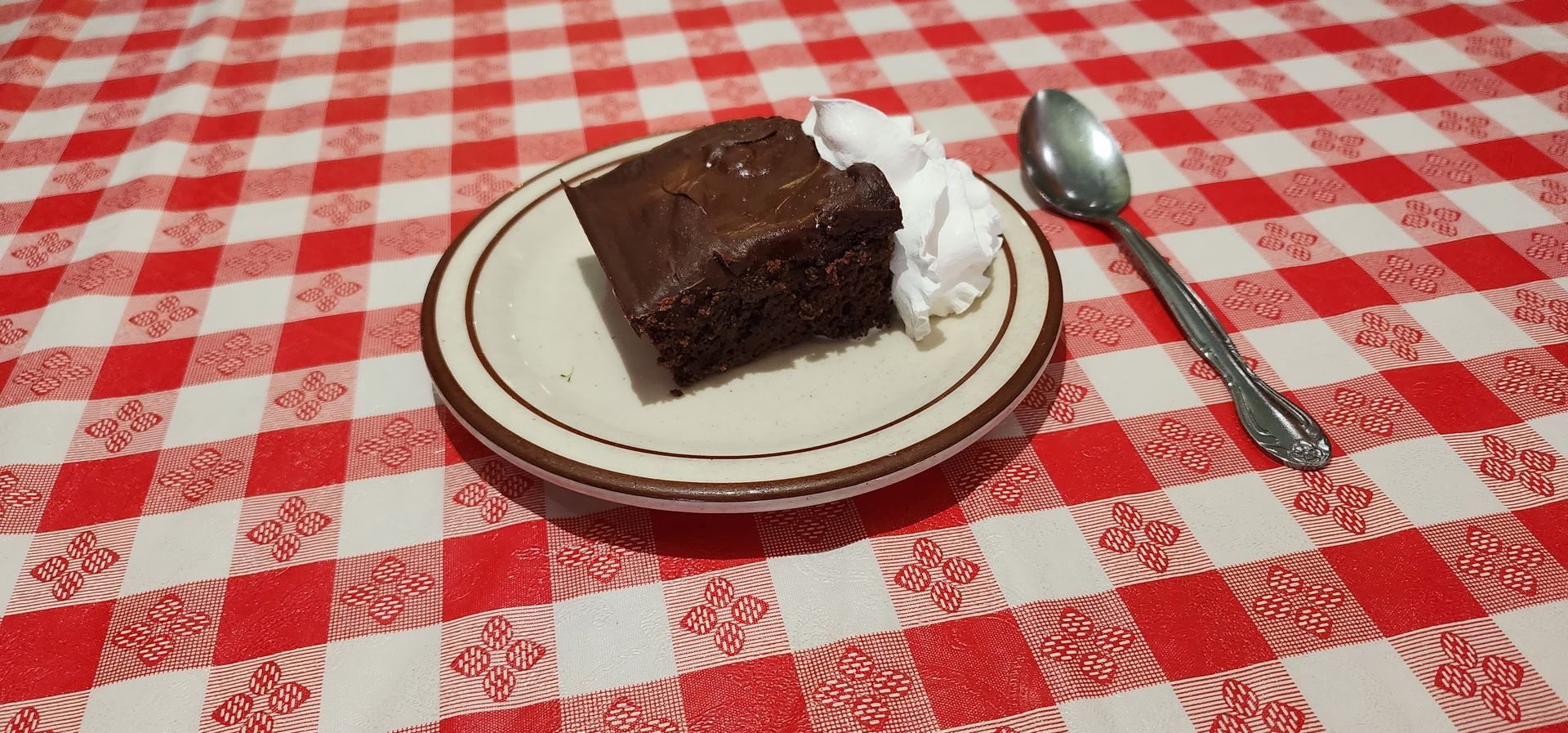A piece of chocolate cake on a plate with whipped cream and a spoon on a checkered table cloth.