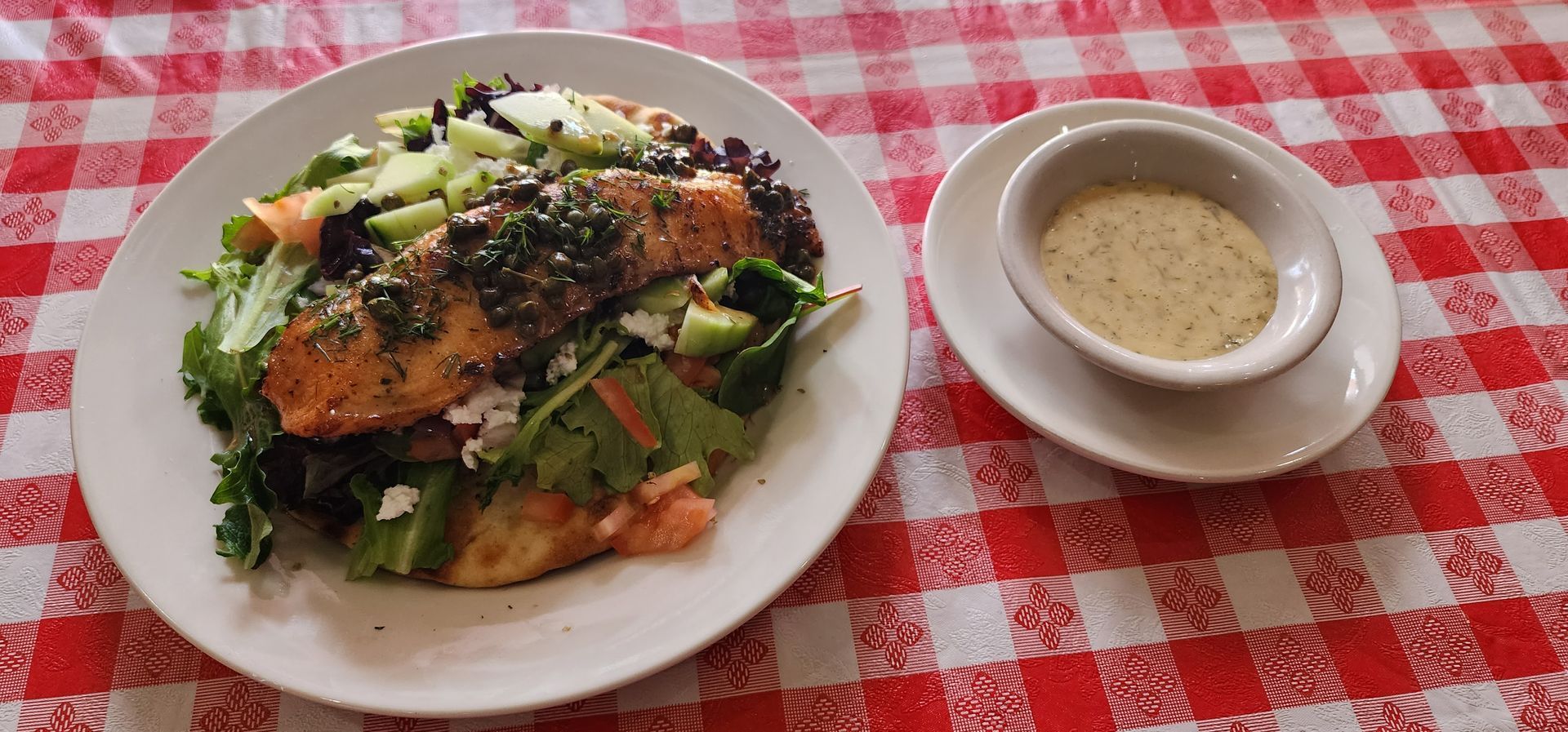 A plate of food with a bowl of dressing on a checkered table cloth.