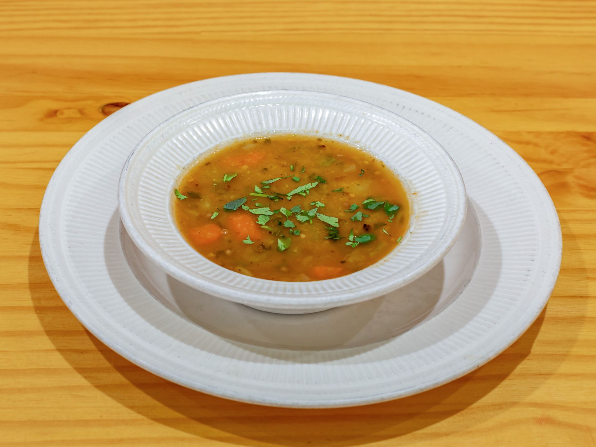 A bowl of soup is on a white plate on a wooden table.