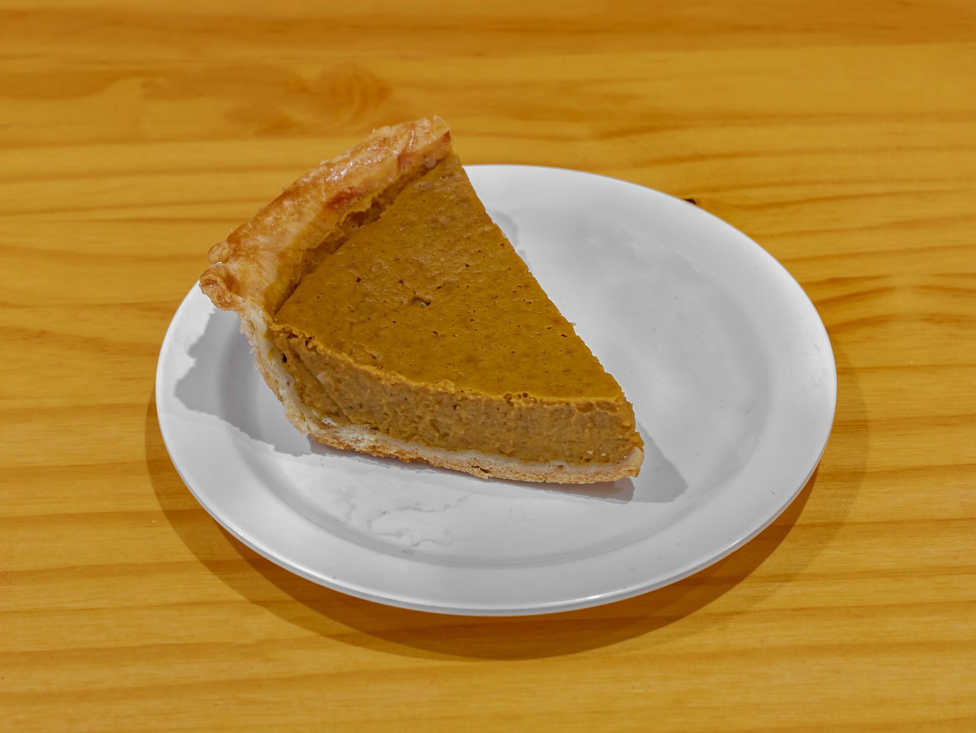 A slice of pumpkin pie is on a white plate on a wooden table.