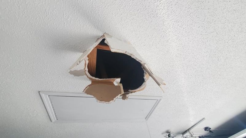 A large, jagged hole in a white textured ceiling located just above a rectangular attic access panel.