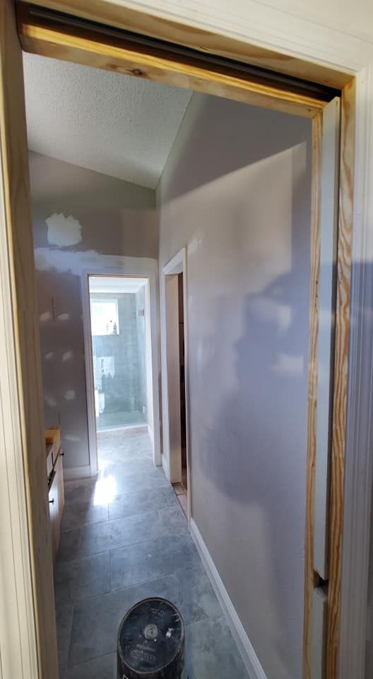 An interior view looking through a wooden door frame into a hallway with grey tiled floors, white walls, and a bathroom.