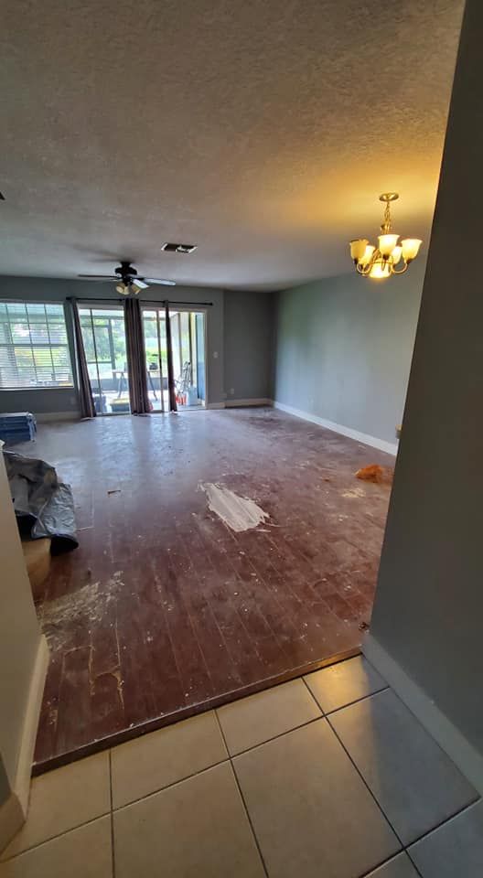 A view from a tiled entryway into a large, unfinished room with a worn wooden floor, a ceiling fan, and a chandelier.