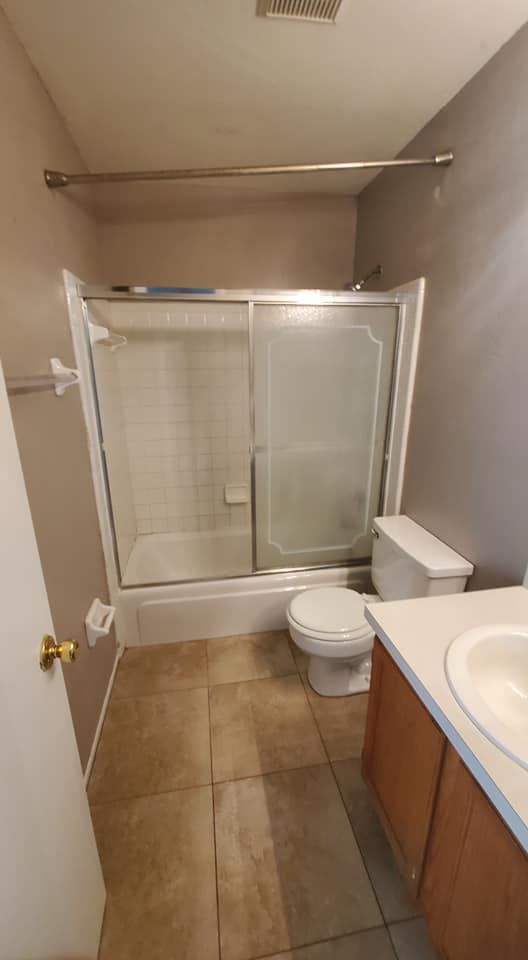 A bathroom with tan tile flooring, a shower-tub combo with a sliding glass door, a white toilet, and a vanity sink.