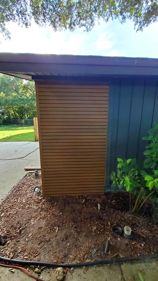A house exterior wall featuring horizontal brown wood slats adjacent to dark vertical siding, set above a mulched garden.