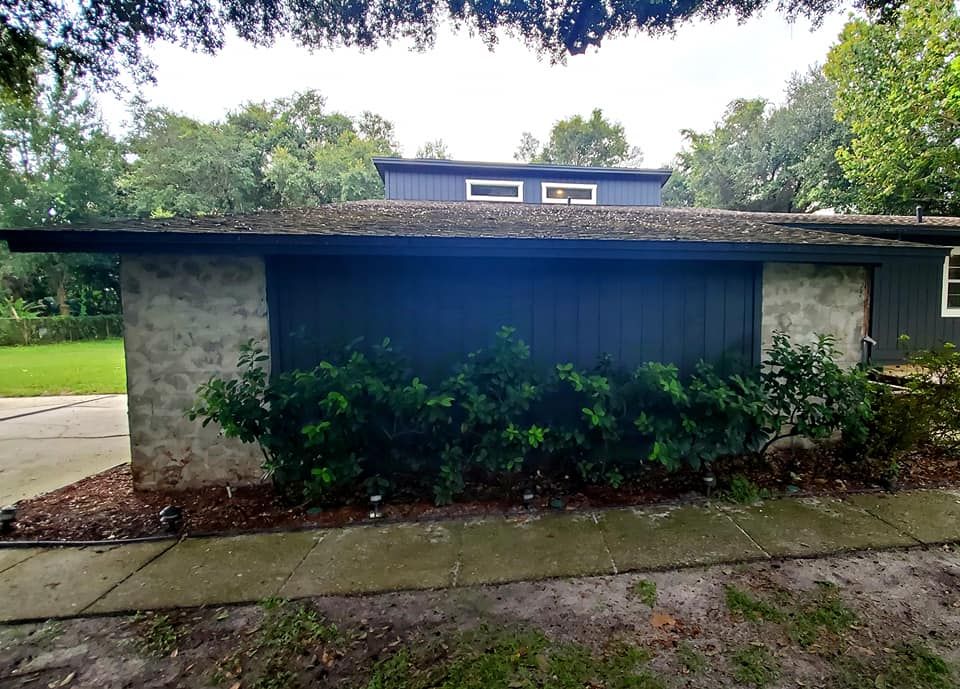 A mid-century home exterior with a dark blue vertical-siding wall, stone corners, a shingled roof, and a row of shrubs.