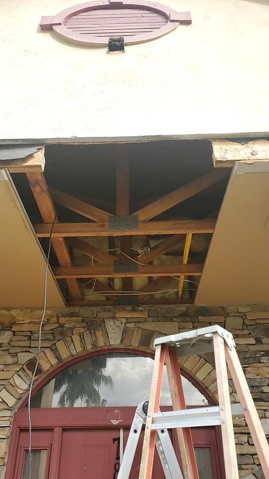 A ladder stands below an exterior ceiling section where plaster has been removed, revealing wooden roof framing and beams.