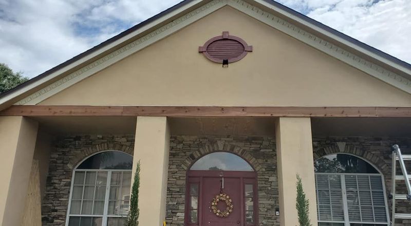 The front exterior of a beige house featuring a stone facade, arched windows, a red front door, and a horizontal beam.