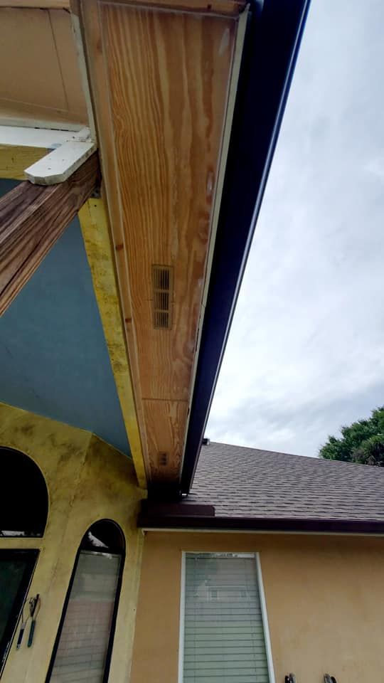 Low-angle view of a house exterior showing exposed wood soffit with vents, a brown shingled roof, and tan-painted walls.