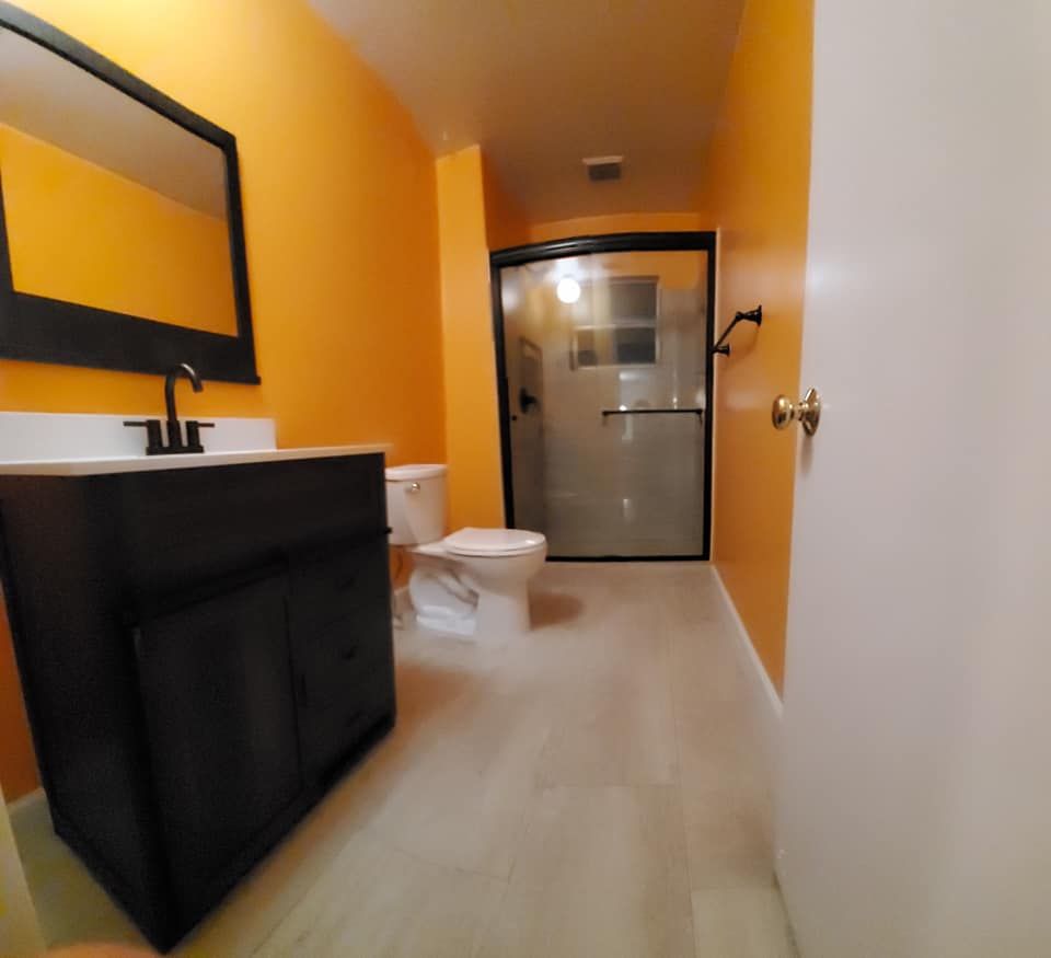 A bathroom with bright orange walls, a black vanity, a rectangular mirror, a white toilet, and a glass-enclosed shower.