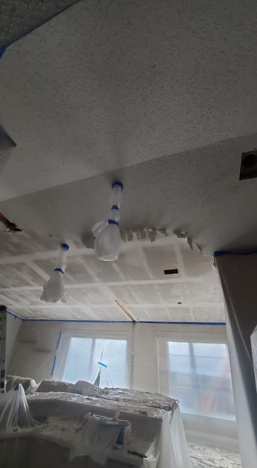 A room under renovation with ceiling drywall taped and sanded, plastic-covered light fixtures, and windows masked off.