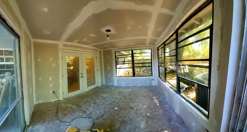 A sunroom interior under renovation featuring unpainted drywall, a central ceiling light, and large windows.