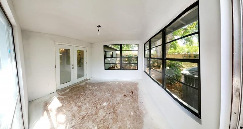 A sunroom with white walls, French doors, black-framed windows, and a floor covered in construction debris.
