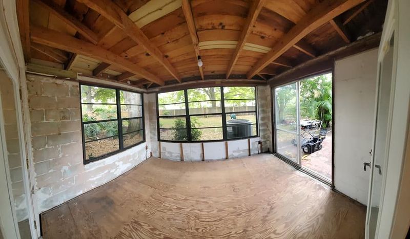 A sunroom under renovation featuring exposed wood ceiling beams, unfinished concrete block walls, and a plywood floor.