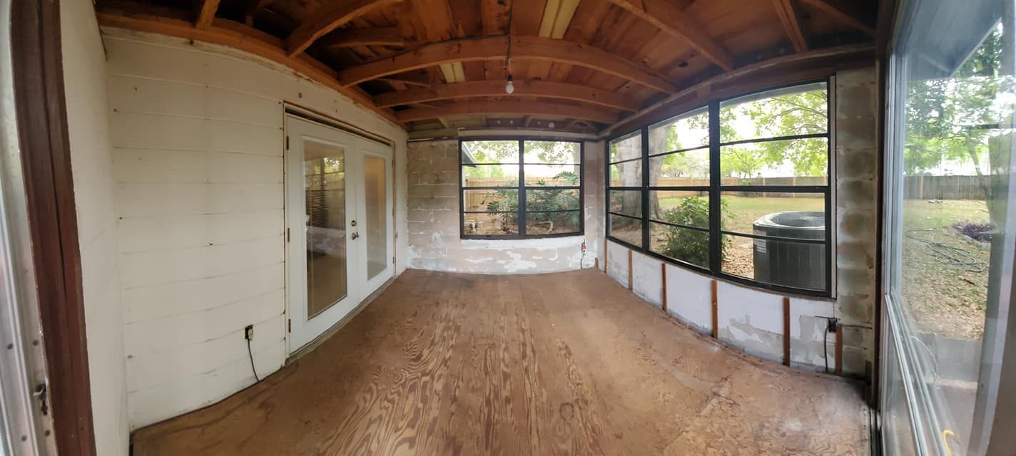 A wide-angle view of a sunroom under construction, featuring exposed wooden ceiling beams, plywood flooring, and windows.