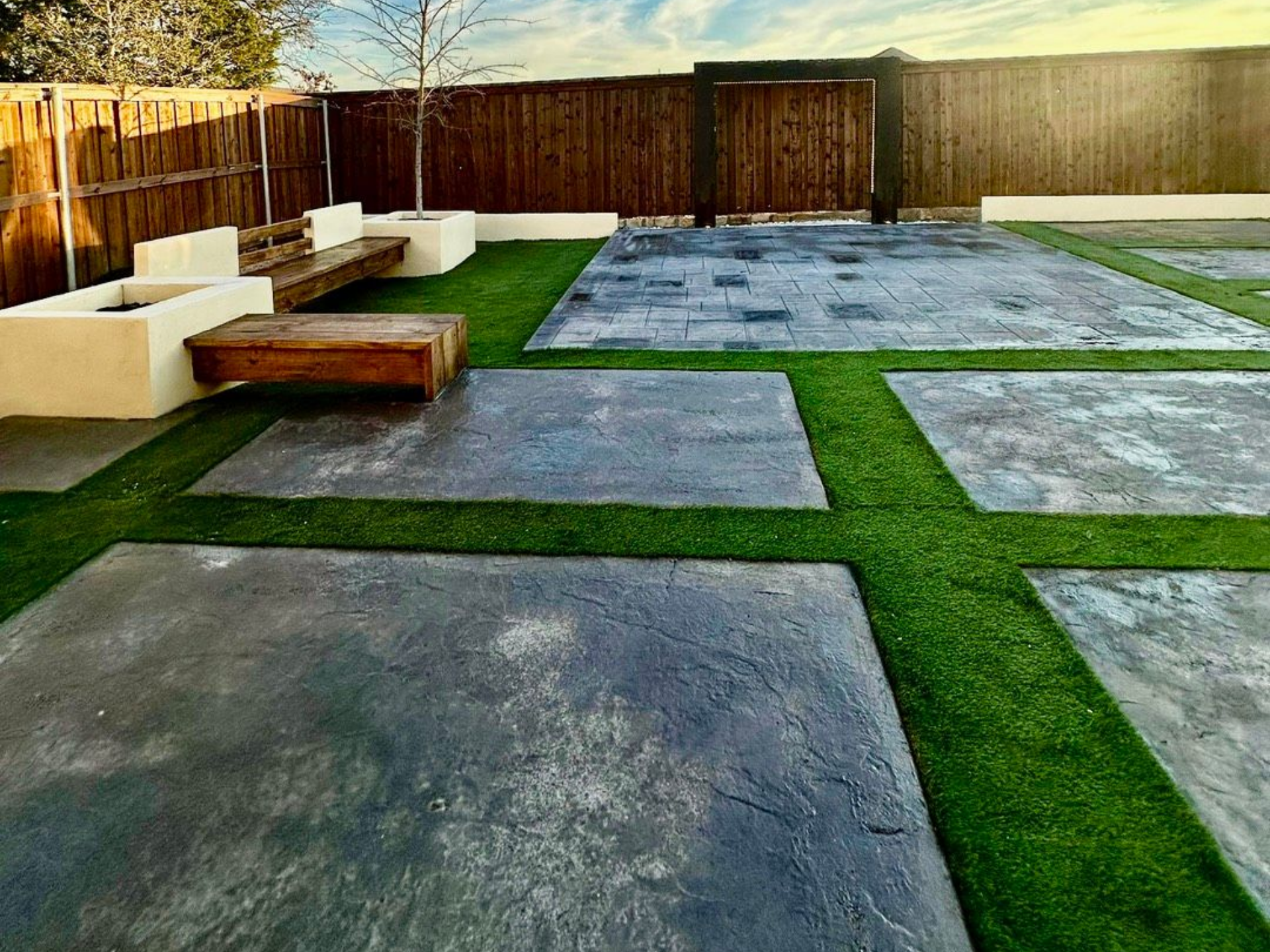 Backyard patio with square stone pavers and artificial grass, wooden bench, and wooden fence under a blue sky.