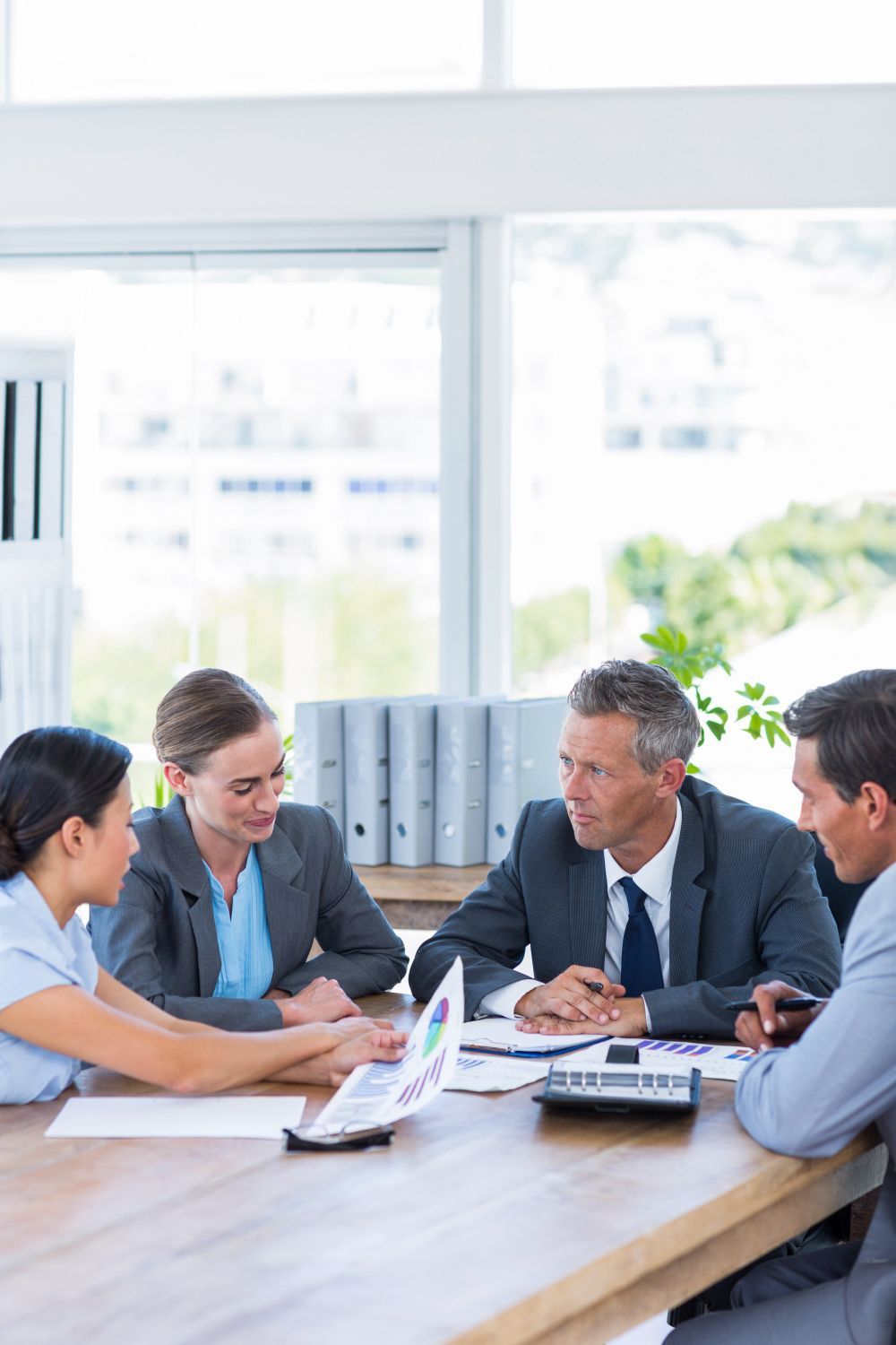 Businesspeople in suits around a table, reviewing documents and discussing strategy in an office.