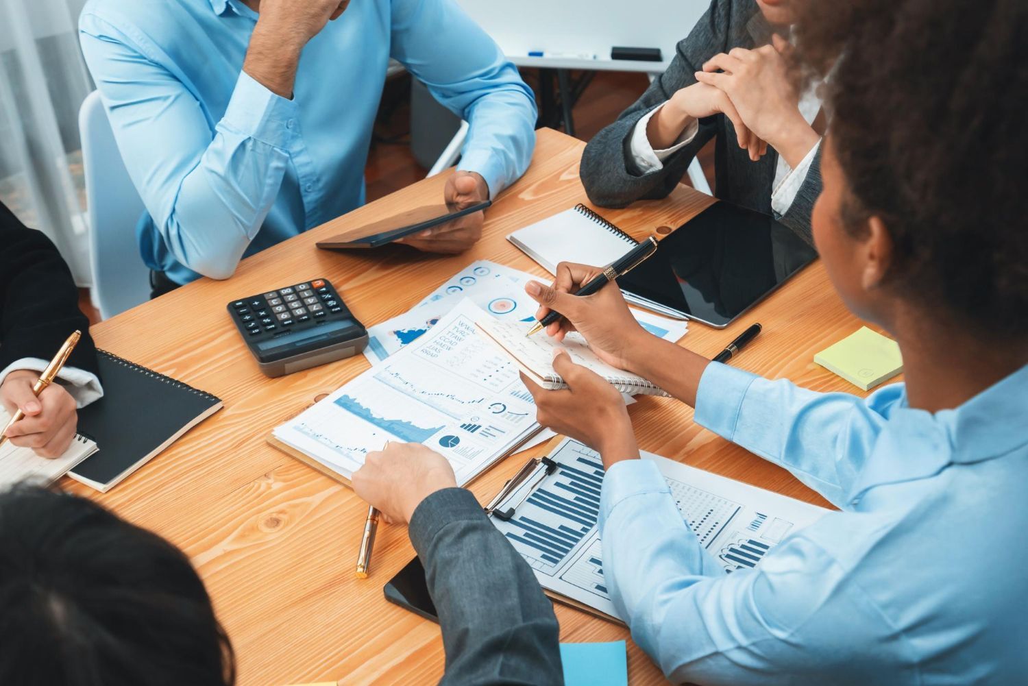 People in business attire at a wooden table reviewing financial data; using calculator, tablet and pens.