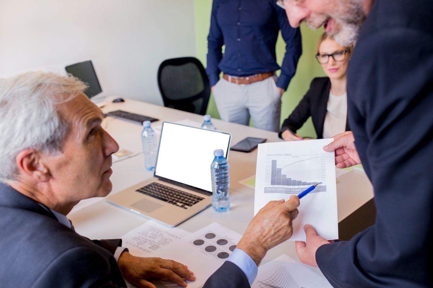 Businesspeople in a meeting, reviewing documents and a bar graph. A laptop and water bottles on the table.
