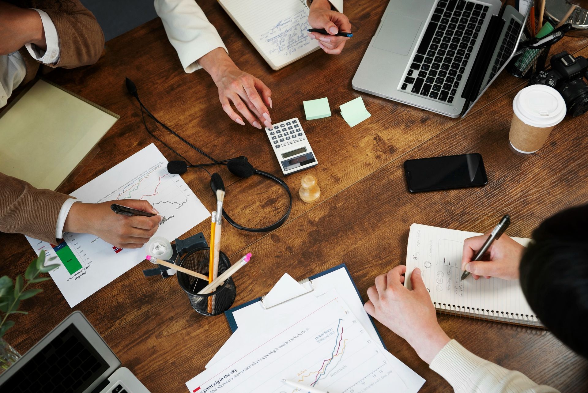 People at a wooden table collaborating with papers, calculator, laptop, phone, and coffee cup.