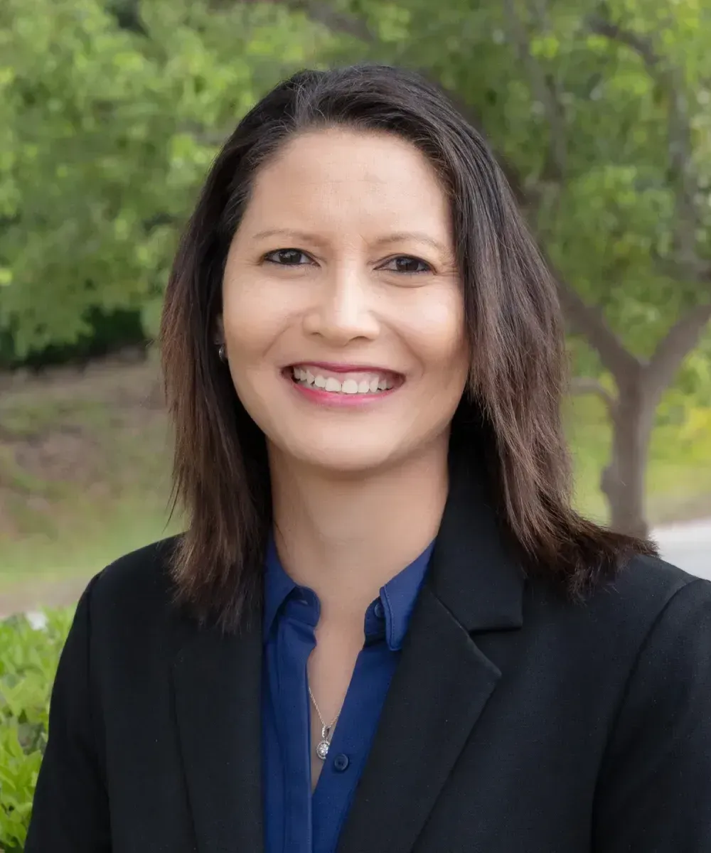 Woman in gray blazer smiles outdoors with blurred greenery.