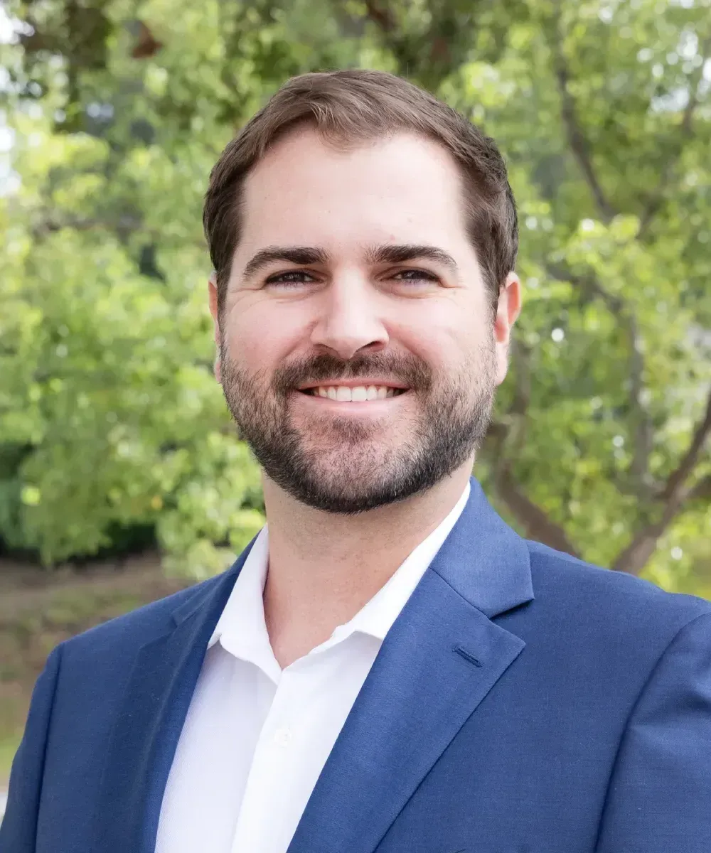 Man in suit and patterned shirt smiles in front of green foliage.