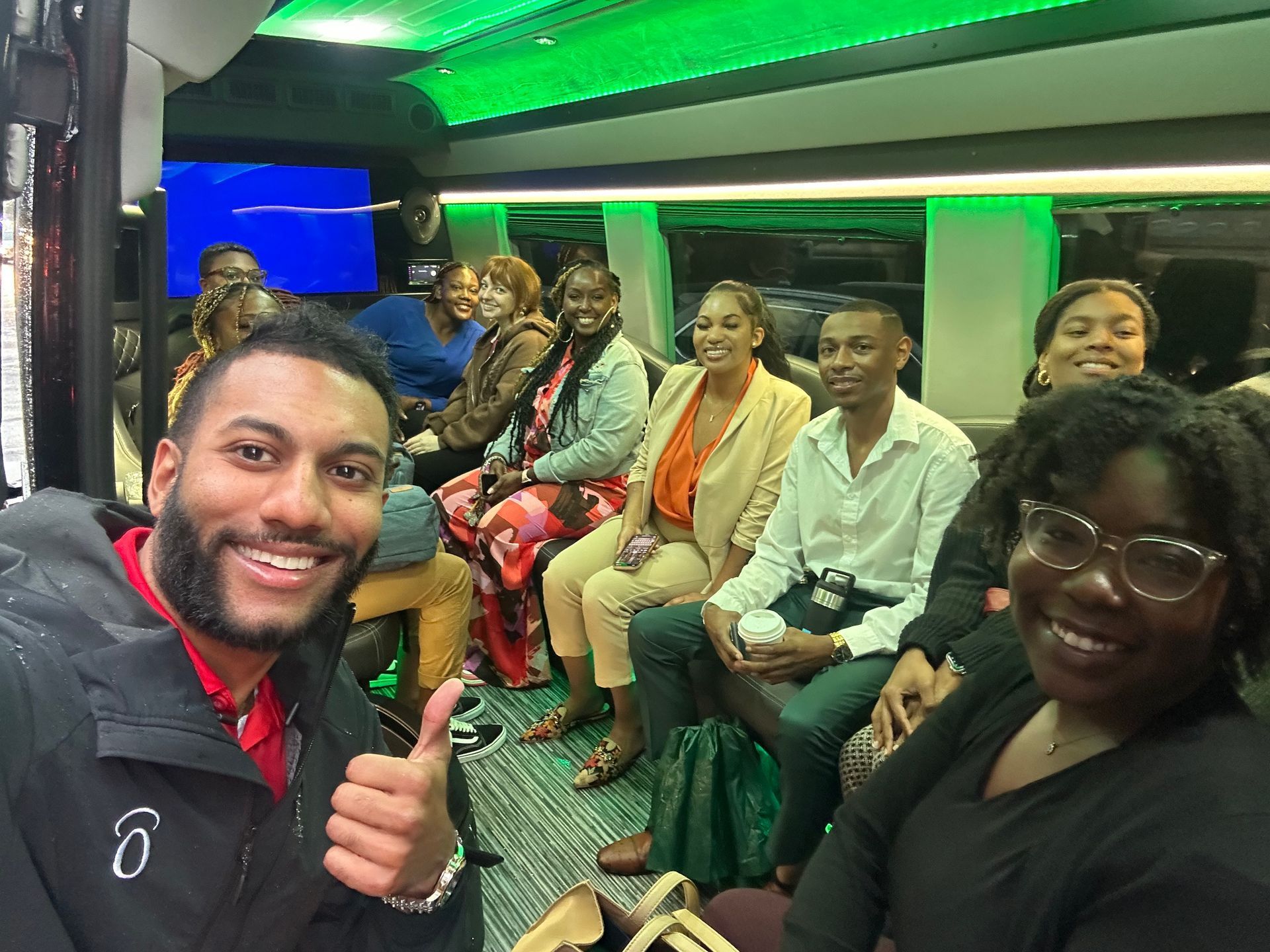 Group of people smiling inside a vehicle with green lighting, one giving a thumbs-up.